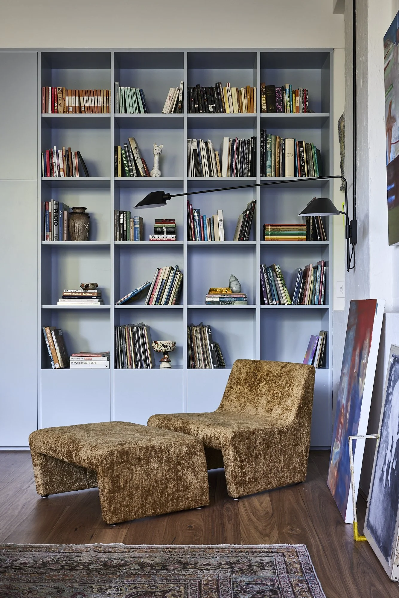 A blue bookshelf filled with books, decorative items, and art pieces, with a brown upholstered chair and ottoman in front of it, on a wooden floor with a rug to the side.