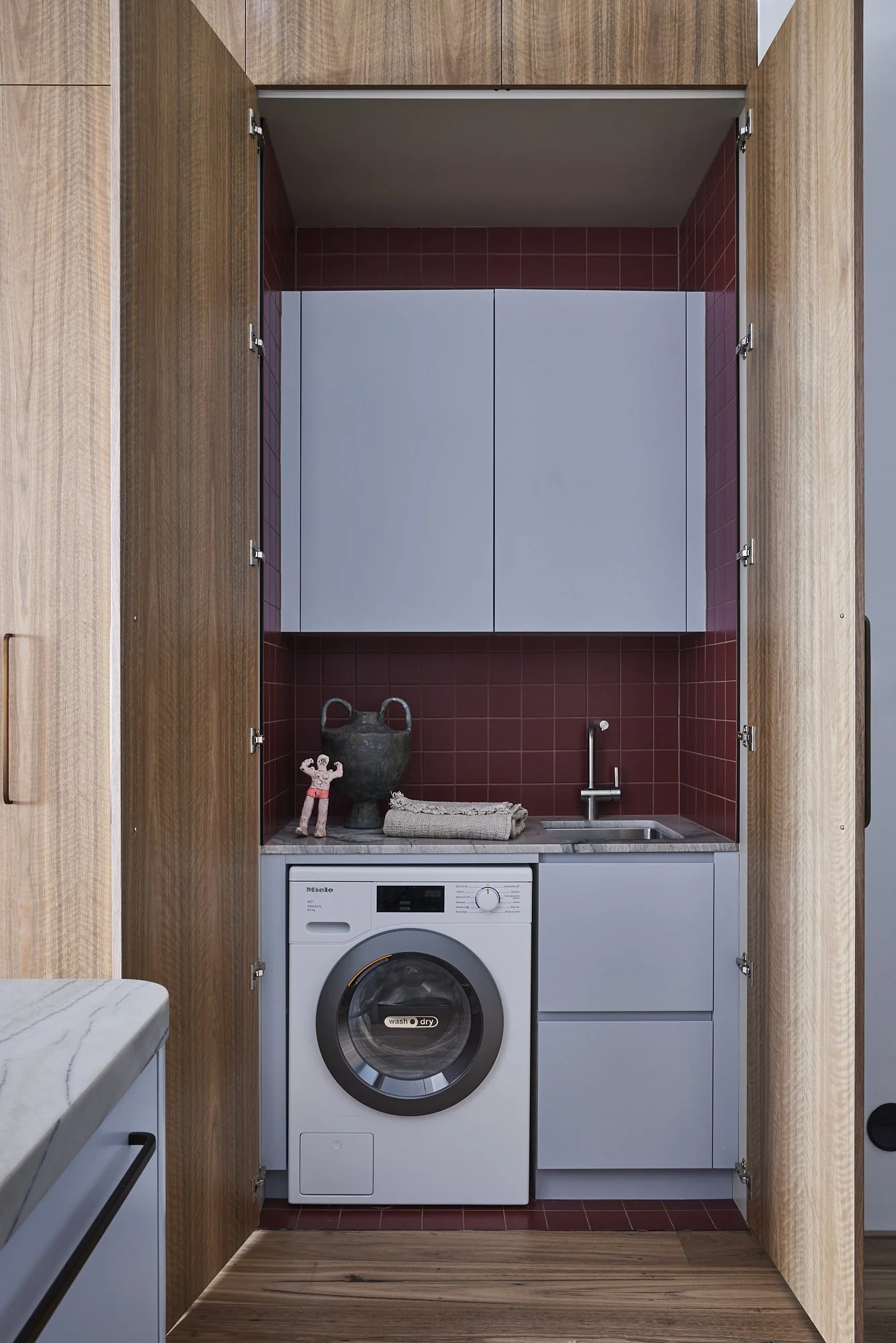 A compact laundry area inside a wooden cabinet with doors open, featuring a front-loading washing machine, a small sink, a faucet, a countertop with decor including a large vase, a small figurine, and a rolled towel, with blue and red tile accents.