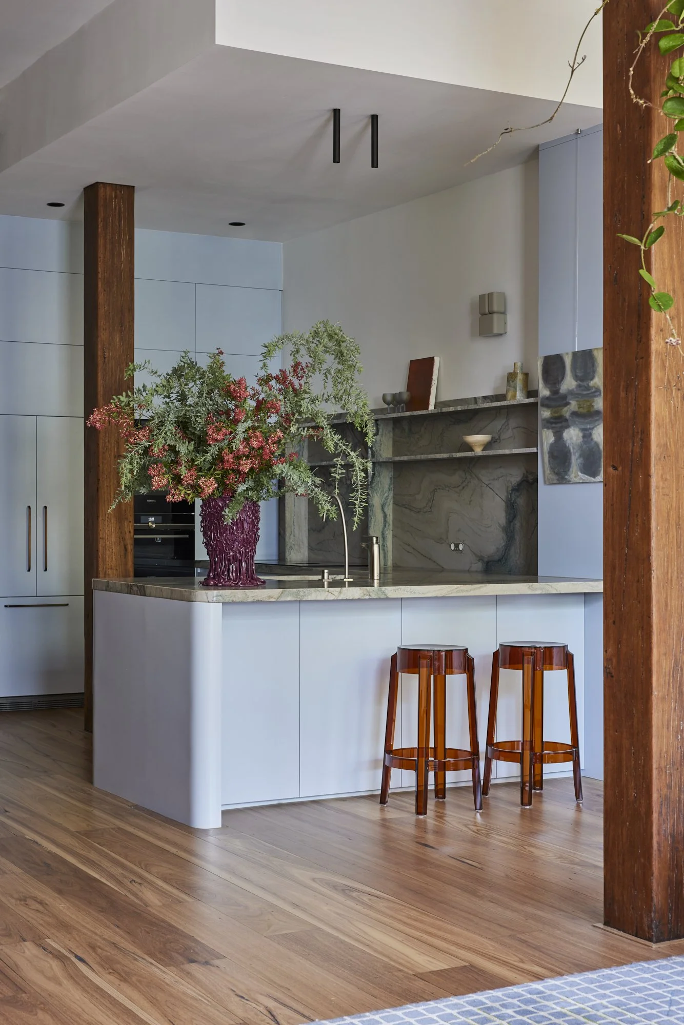 A kitchen with a marble countertop, a large floral arrangement in a purple vase, and two orange-red translucent bar stools.