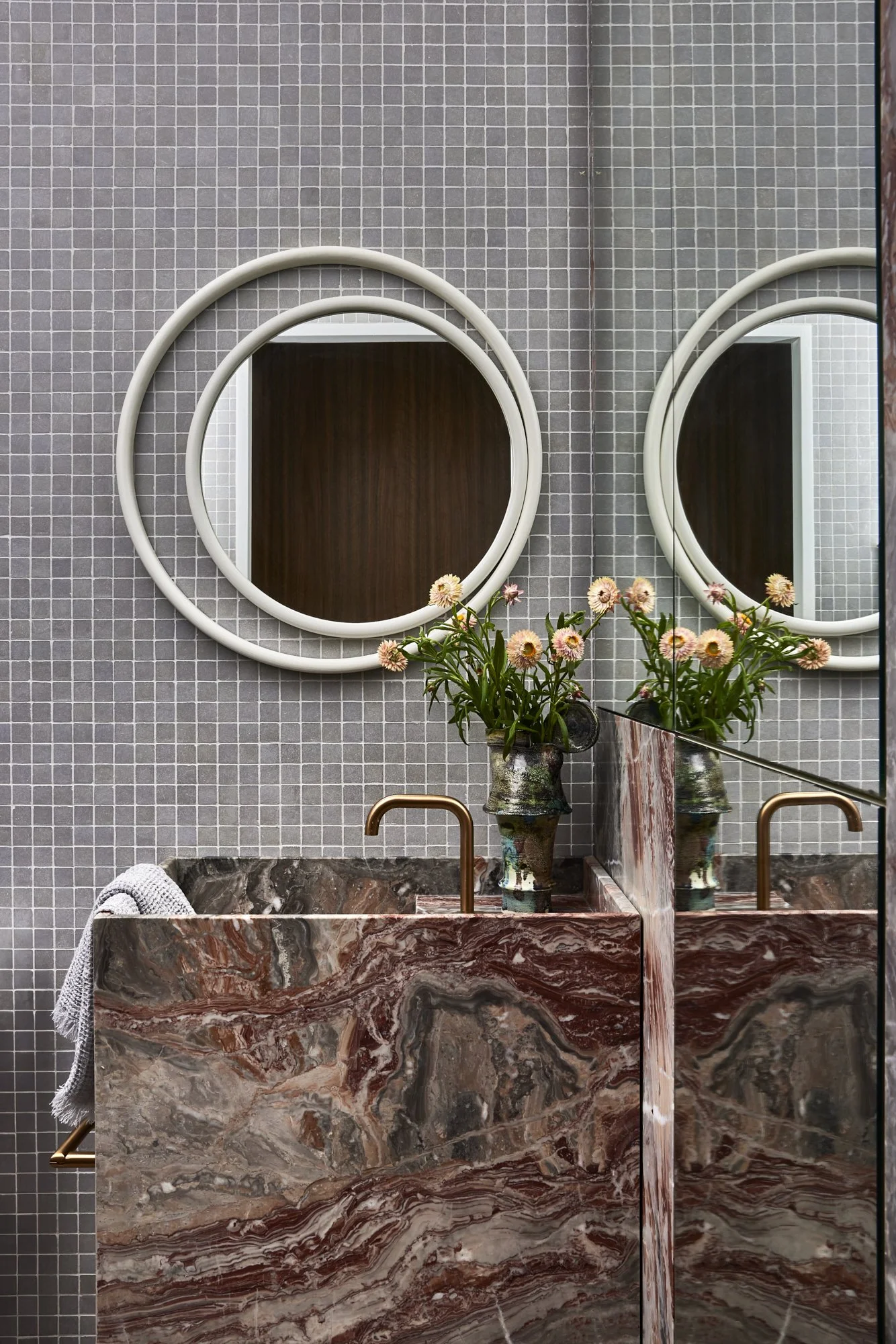 A bathroom with gray tiled walls, a marble sink with red, gray, and white patterns, two oval mirrors above the sink, two gold-colored faucets, a vase with pink flowers on the sink, and a cloth hanging on the left side.