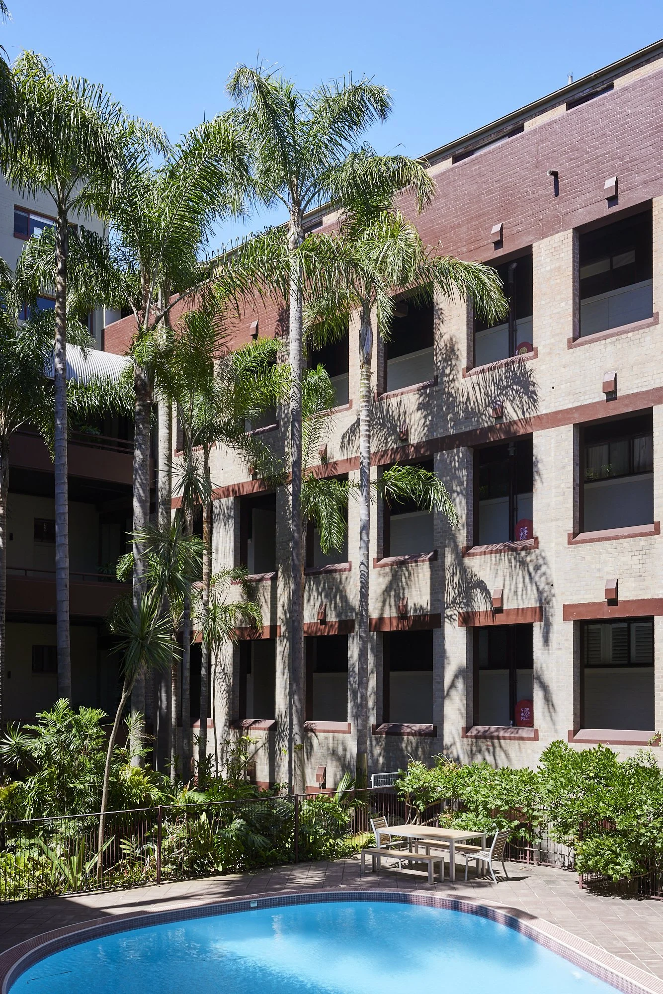 A swimming pool with a fenced garden area, surrounded by tall palm trees and multi-story brick apartment building with open windows under a clear blue sky.
