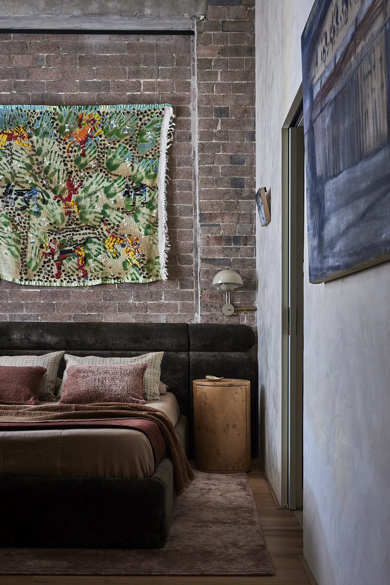 A bedroom with a dark upholstered bed, pink and neutral pillows, and a brown bedspread. Behind the bed is an exposed brick wall with colorful, patterned textile art hanging on it. To the right, a white wall features a modern bent-arm wall lamp. There is a small wooden side table next to the bed and a large abstract painting on the white wall.