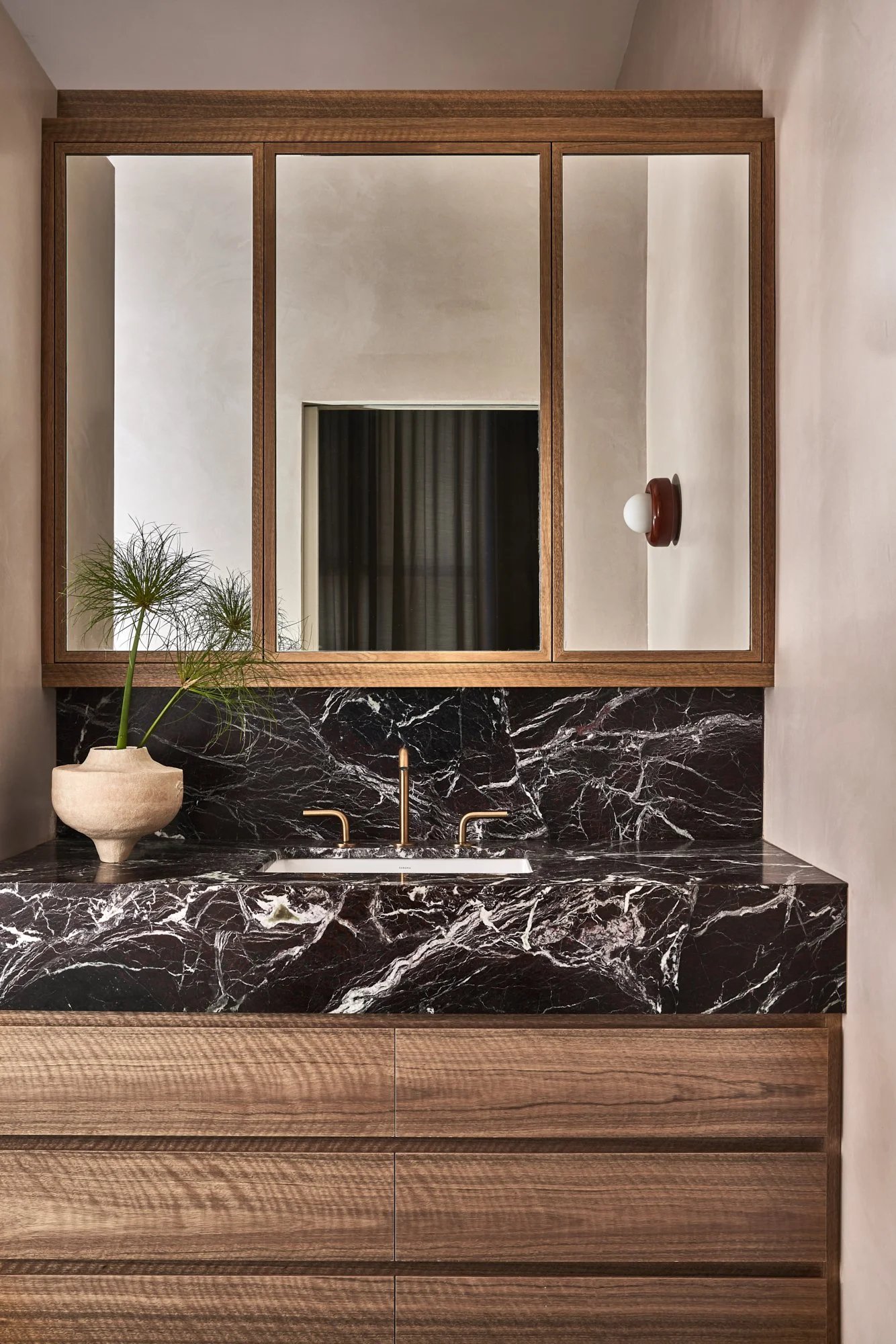 A bathroom vanity with a black marble countertop, a small white sink, and gold fixtures. There is a large wooden framed mirror above the sink, and a potted plant on the left side of the countertop.