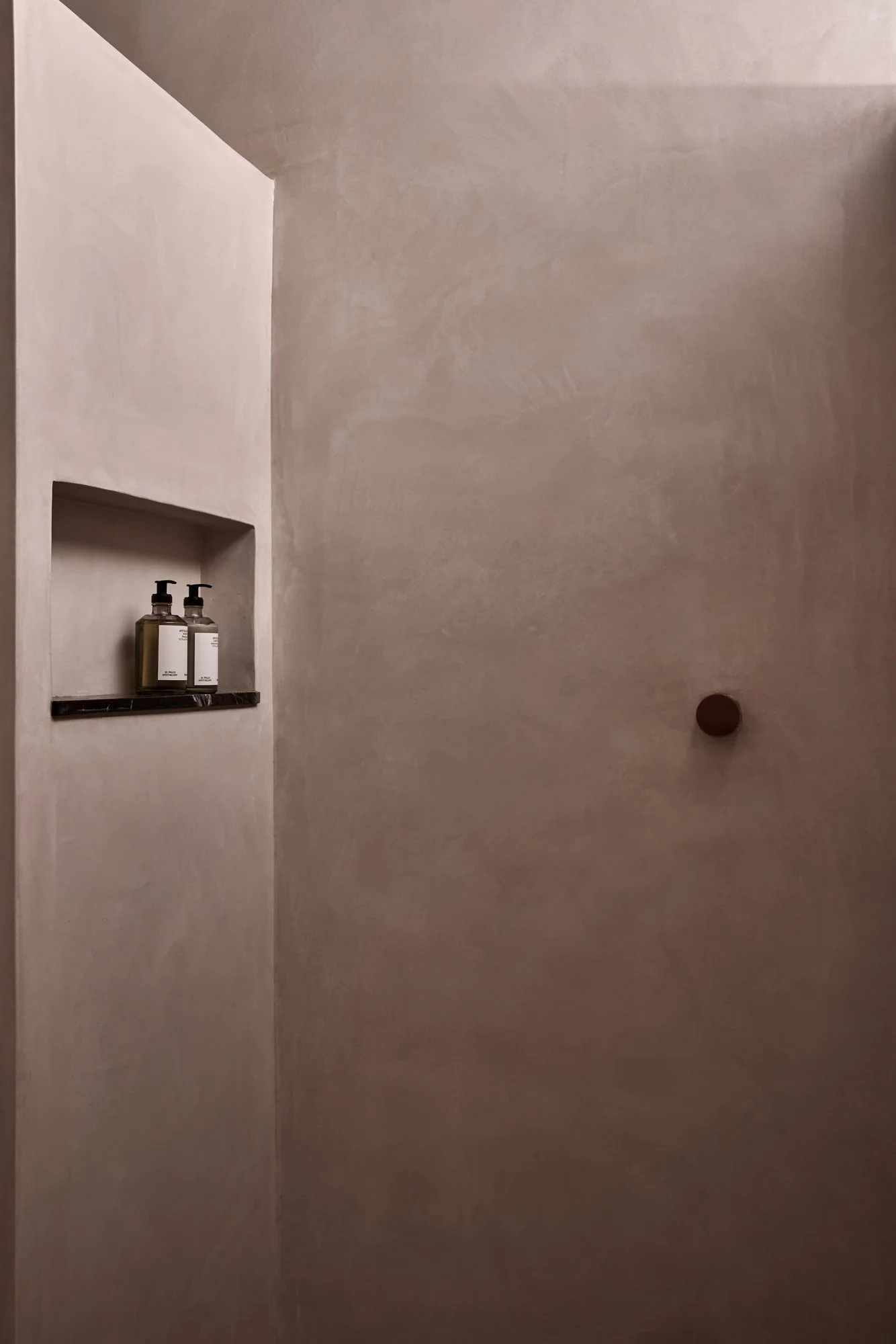 Minimalist shower with two bottles of body wash and a soap dispenser on a black shelf, with a round brown handle on the door.