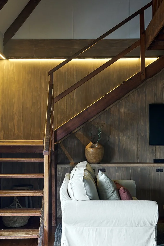 Interior living space with wooden staircase, beige sofa with pillows, potted plant on a shelf, and a wall-mounted TV