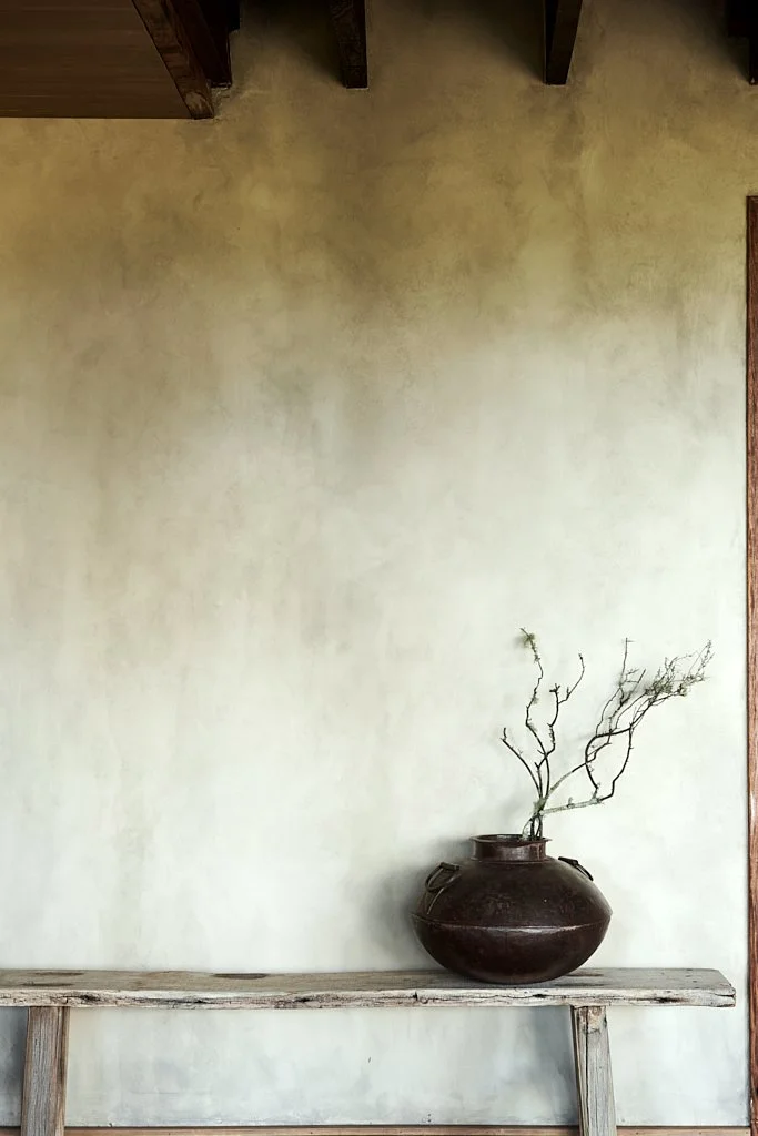 A minimalist interior with a rustic wooden bench and a large dark brown vase holding twigs, against a textured light beige wall.