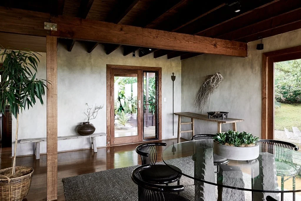 A dining area with a round glass table surrounded by black chairs, a potted plant on the table, a large window to the outside, a wood door leading outdoors, and minimalist decor with plants and a light-colored wall.