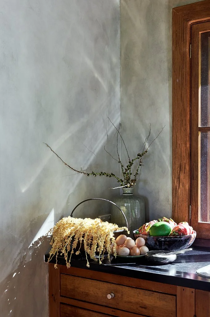 A kitchen corner with a neutral wall, a wooden cabinet, and a black countertop. On the counter, there is a clear glass vase with bare, twisted branches, a metal basket with a bunch of beige dried flowers, a bowl of eggs, and a bowl with colorful decorative eggs and roses.