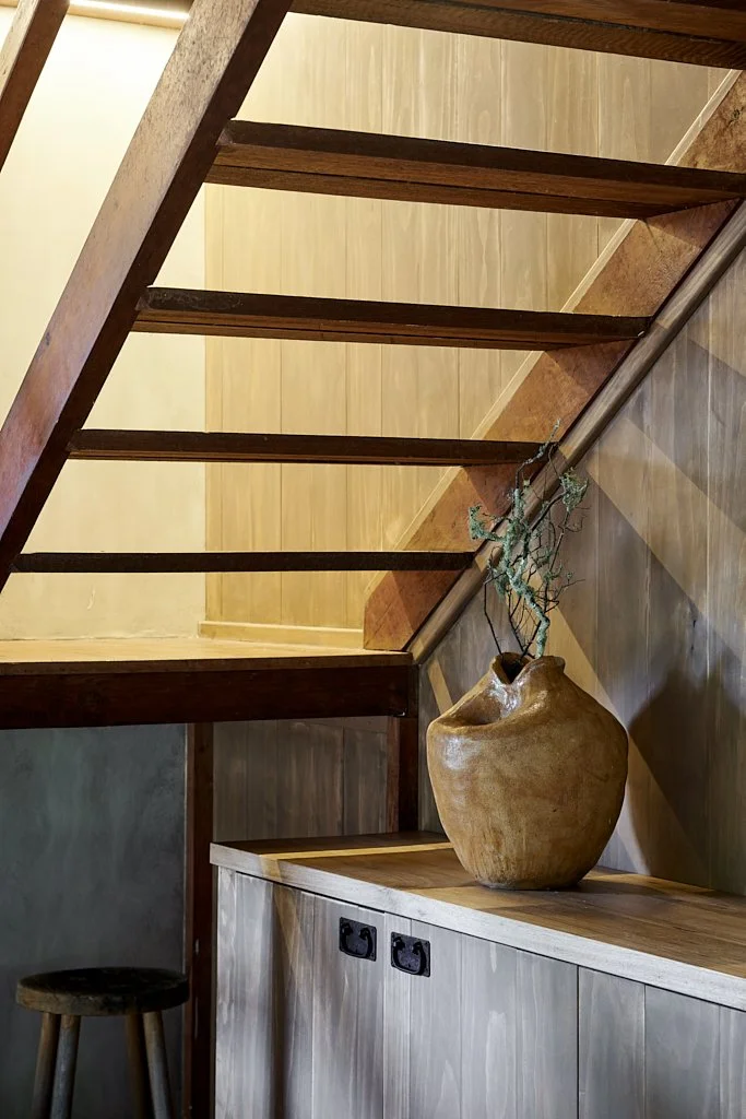 Interior view of a staircase with wooden steps, a wooden handrail, a wooden cabinet with black electrical outlets, a decorative vase with a plant, and a stool in the corner.