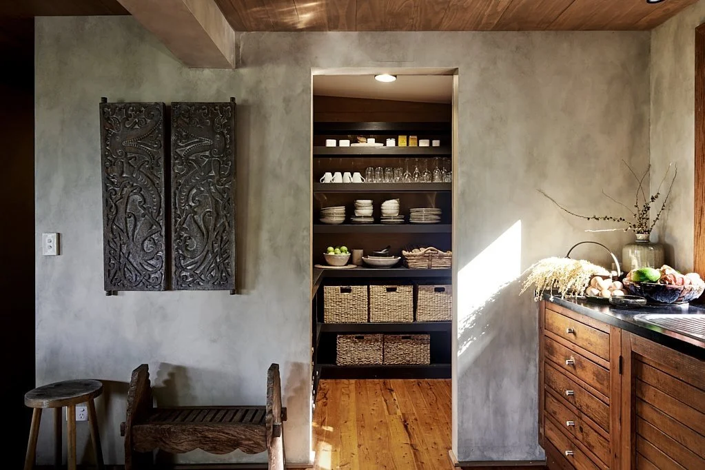 Interior of a kitchen with wooden cabinetry on the right, open shelving in the center with dishes, glasses, and baskets, and a decorative wooden wall hanging on the left. Natural light illuminates part of the space.