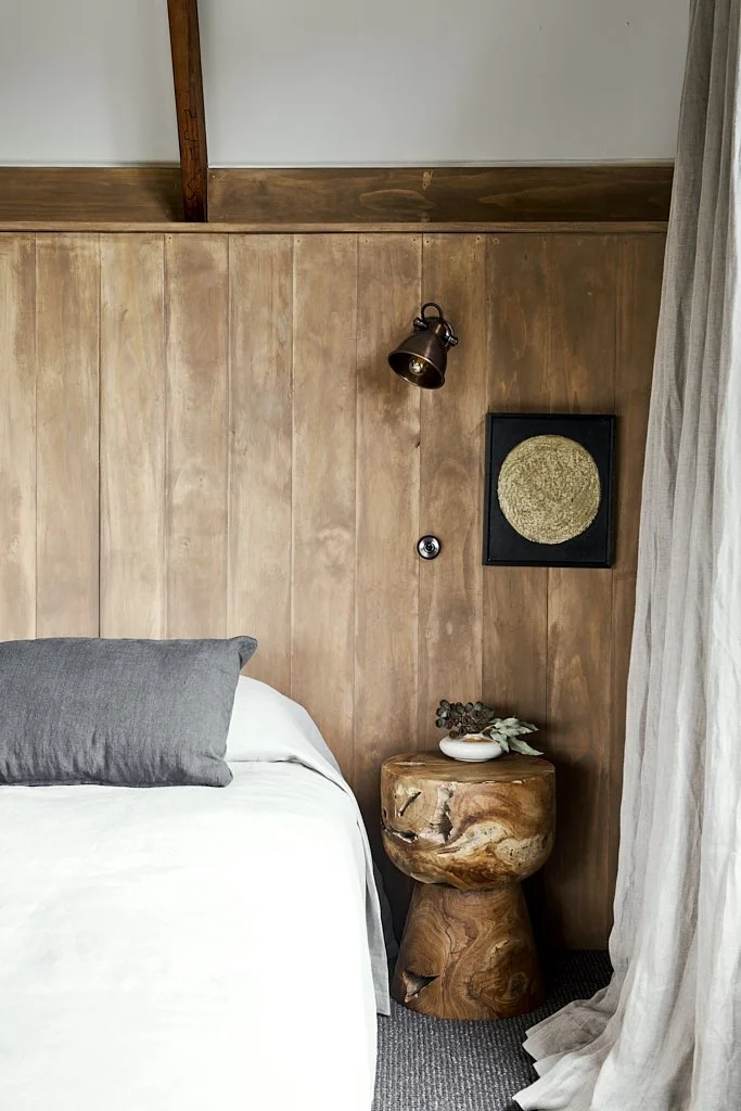 A cozy bedroom corner with a bed, a wooden headboard, a gray pillow, a rustic wooden side table with decorative items, a black wall-mounted lamp, a framed circular golden artwork, and a curtain on the right side.