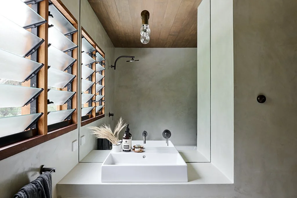 Modern bathroom with a white rectangular sink, wooden window shutters, and a wooden ceiling. The bathroom has a minimalist design with a large mirror reflecting the sink, and a small decorative vase with dried plants on the countertop.