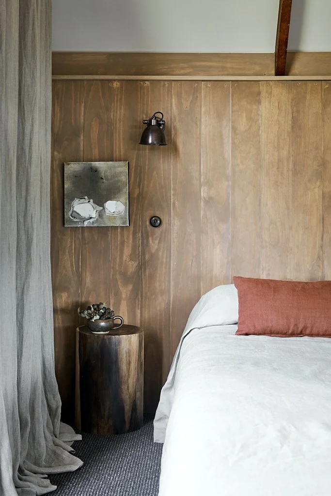 Part of a bedroom with a bed, a rust-colored pillow, a wooden side table with a small plant, wood-paneled wall, a wall-mounted lamp, and a painting on the wall