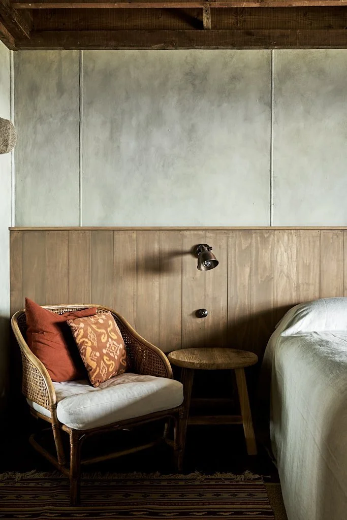 A cozy bedroom corner with a wicker sofa, orange and patterned pillows, a wooden side table, and a bed with white sheets, against a wall with light grey textured paint and wooden paneling, under a wood ceiling.