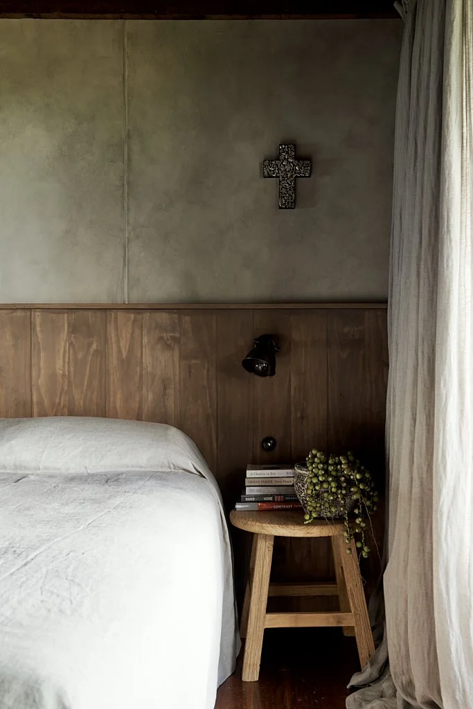 A bedroom corner with a bed, wooden wall paneling, a round wooden side table holding books and a potted plant, a reading wall lamp, and a cross hanging on the wall.