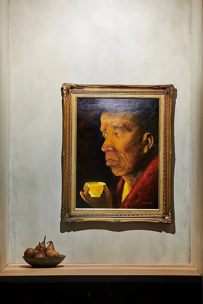 Painting of an elderly woman with Asian features holding a yellow cup. The painting is in a decorative gold frame and is hung on a wall above a shelf with a bowl of pears.