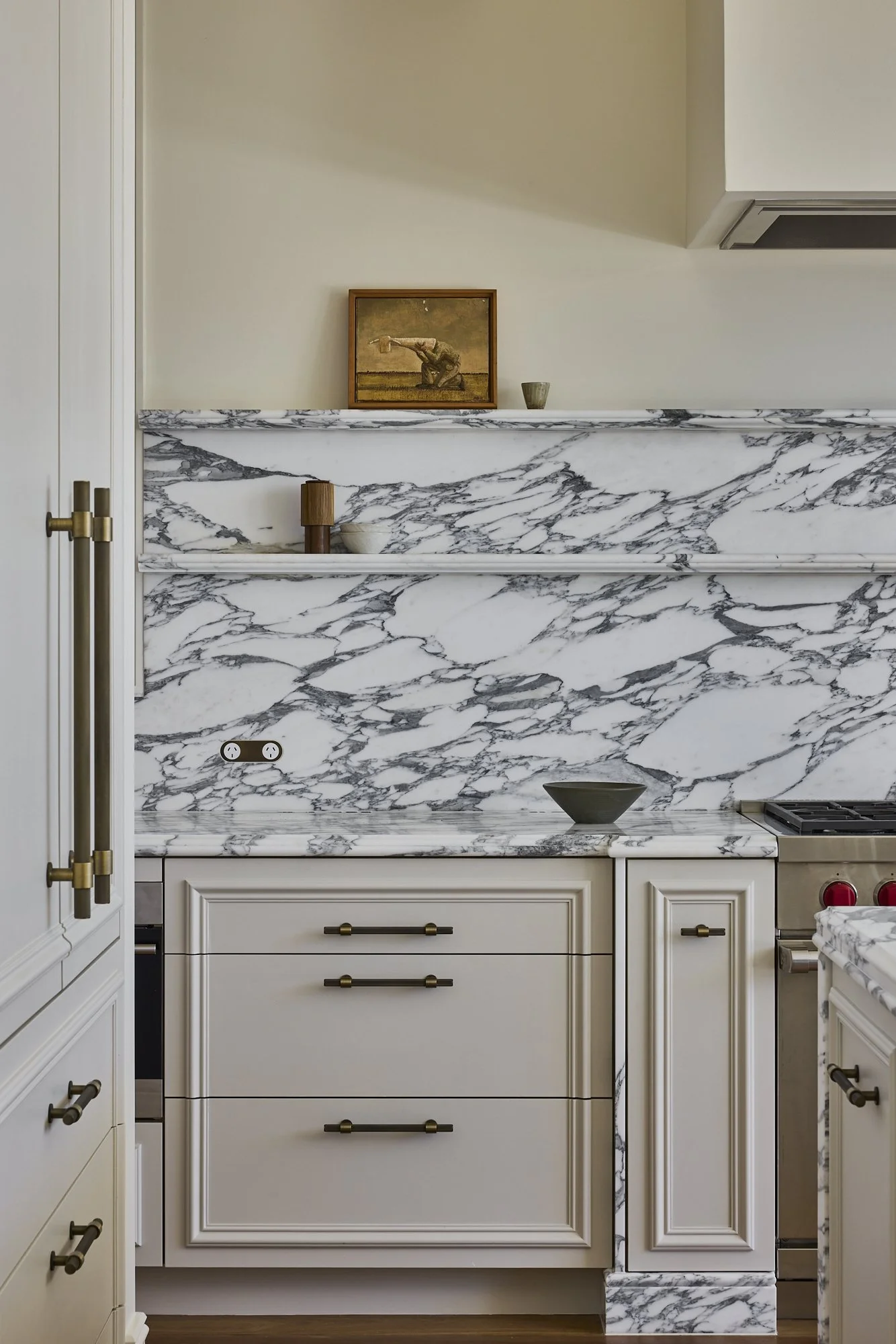 Close-up of a kitchen with white cabinets and a marble backsplash and countertop with gray veining. Decor items include a small bowl, a wooden object, a small pot, and a framed artwork. Parts of a stove and an electrical outlet are visible.