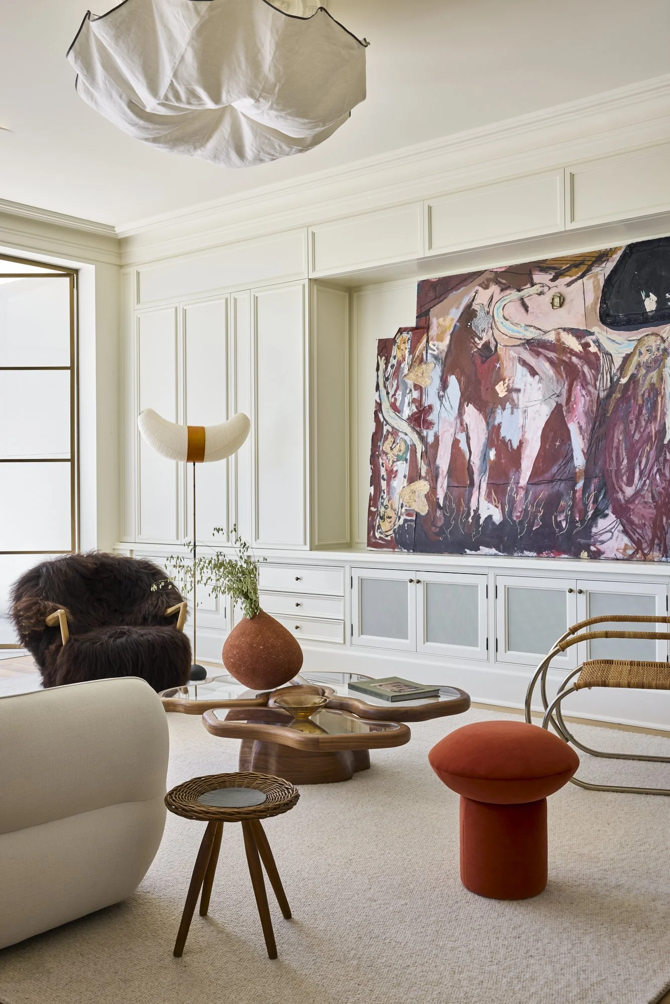 Living room with white built-in cabinets, abstract painting, beige sofa, wooden coffee table, colorful stool, rattan chair, floor lamp, large window, and modern decor.