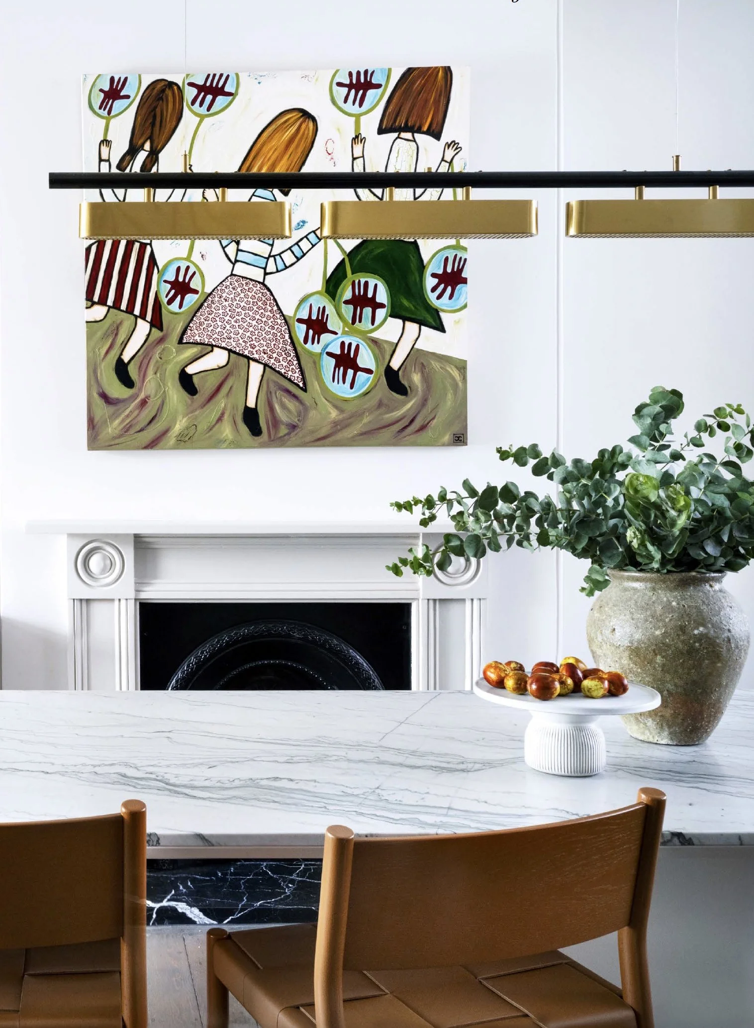 A modern dining area with a white marble table, wooden chairs, a large vase with green foliage, and a white pedestal holding chocolates. Behind the table, there's a white fireplace and a colorful abstract painting of children with long hair and dangling circles, hanging above the fireplace. Gold and black pendant lights are suspended above the table.