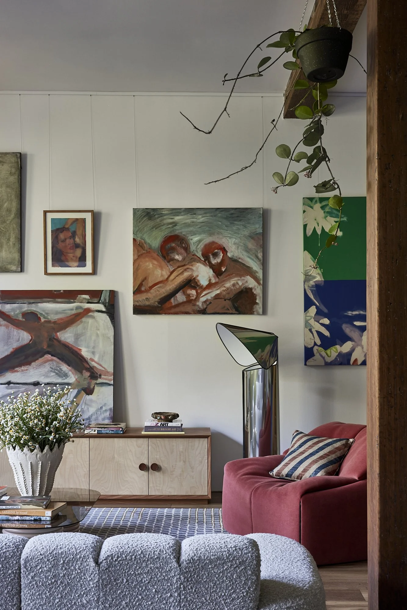 Interior of a living room with a pink armchair, a wooden cabinet, and various colorful paintings on the wall, including a large abstract piece and a floral artwork. A potted plant with trailing vines is hanging from the ceiling.