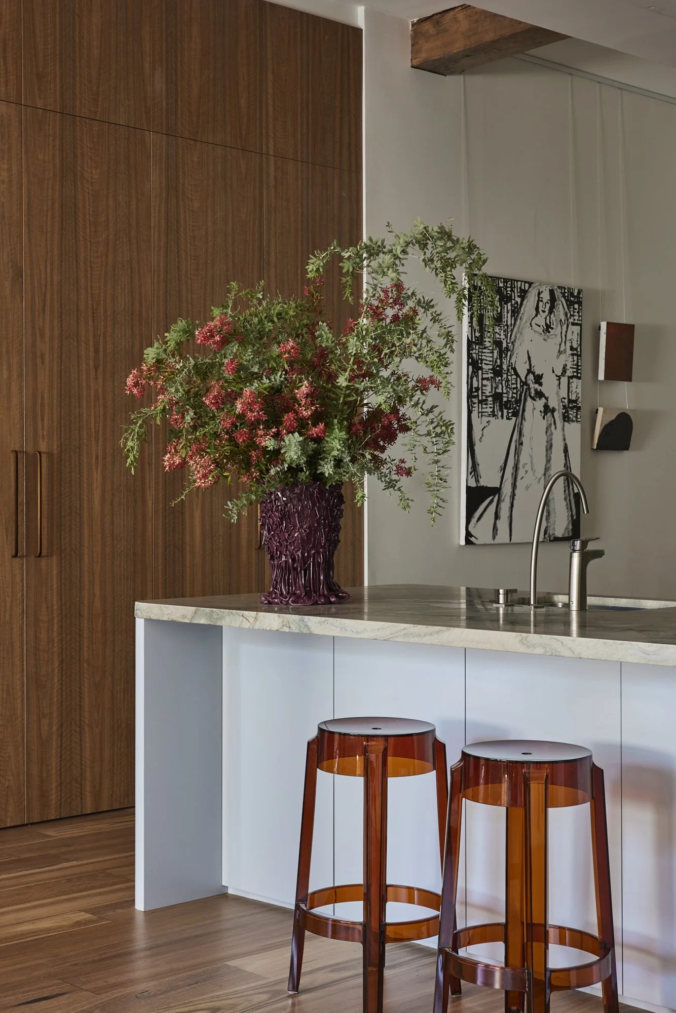A kitchen counter with a large maroon vase filled with pink and green flowers, positioned near wooden cabinets, with two amber-colored transparent bar stools in front, and a black and white artwork on the wall behind the sink.
