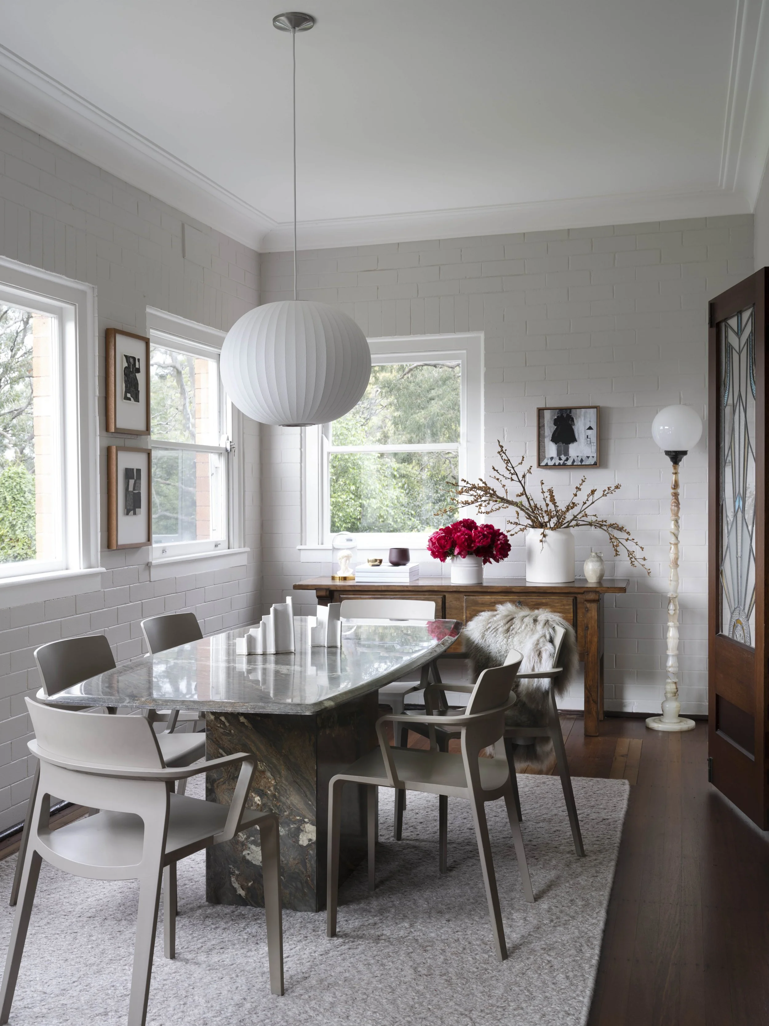 Modern dining room with white walls, large windows, a marble-topped table, six beige chairs, a wooden sideboard with a large vase of pink flowers, framed artwork, a white pendant light, and a tall white floor lamp.