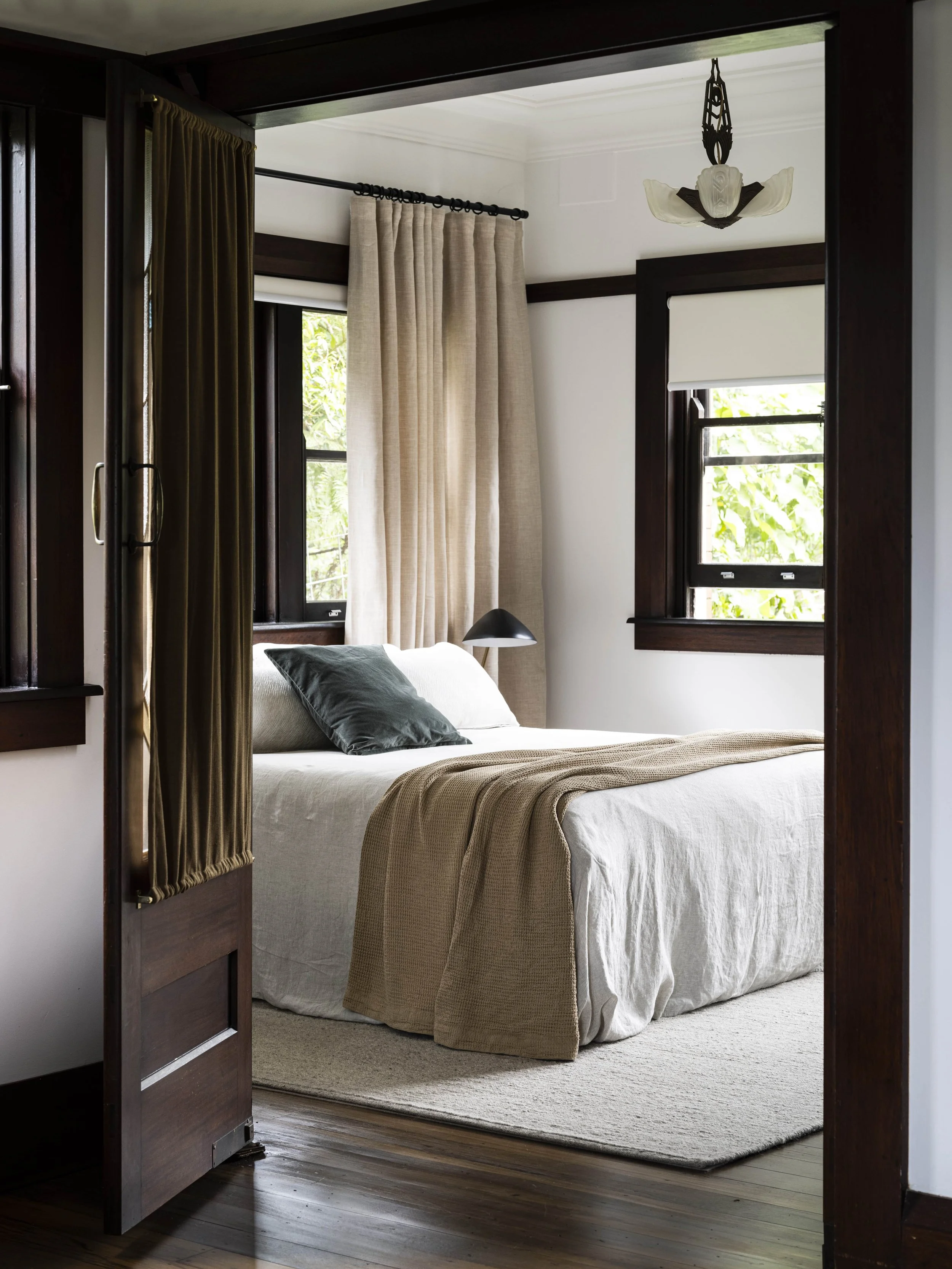 A cozy bedroom with a white bed, dark wooden trim, beige and dark green pillows, a black bedside lamp, and windows with beige and gold curtains, natural light streaming in.