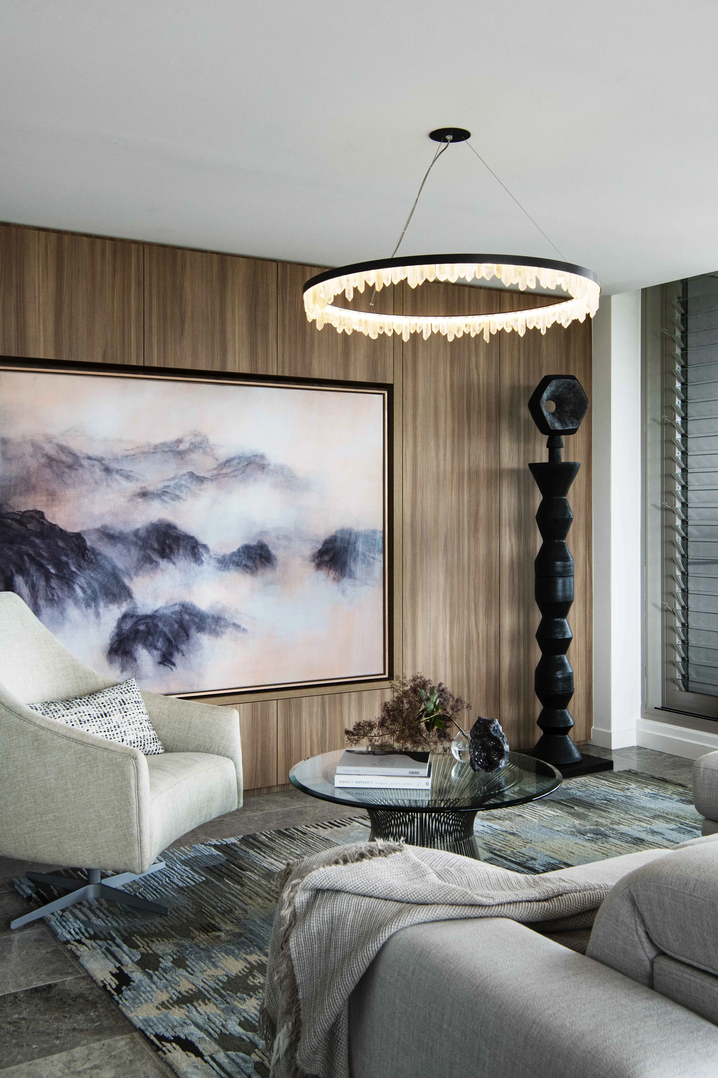 Modern living room with a beige armchair, glass coffee table, large wall art of mountains, wooden wall paneling, oversized black floor lamp, and a chandelier with a black ring and crystal accents.