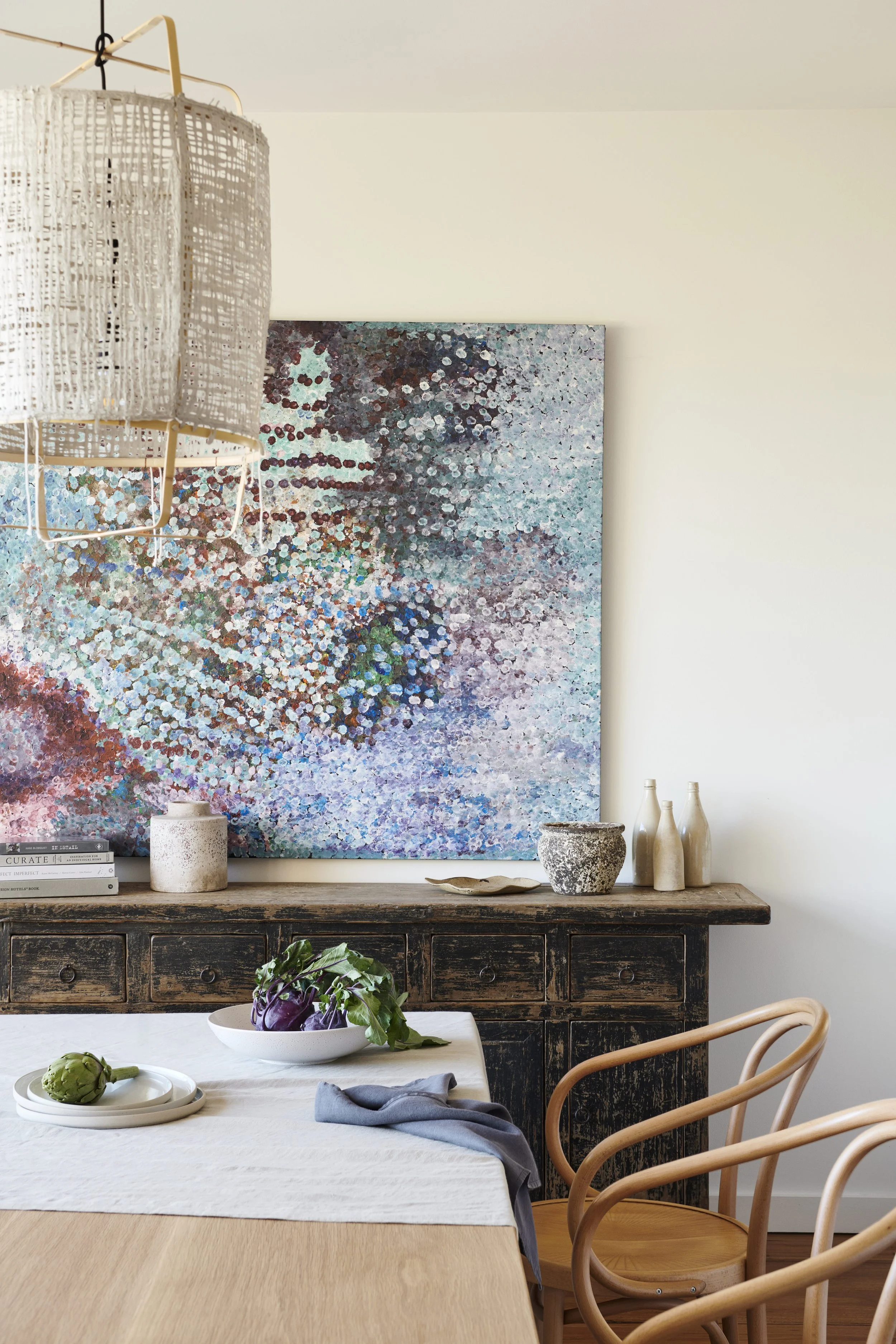 A rustic dining room with a light wood table, a bowl of artichokes, and a white cloth table runner. Two natural wood chairs with curved backs are positioned at the table. Behind, a weathered black wooden sideboard holds books, white vases, and pottery. A large abstract painting with blue, purple, and white colors is hung on the white wall. A woven pendant light hangs from the ceiling.