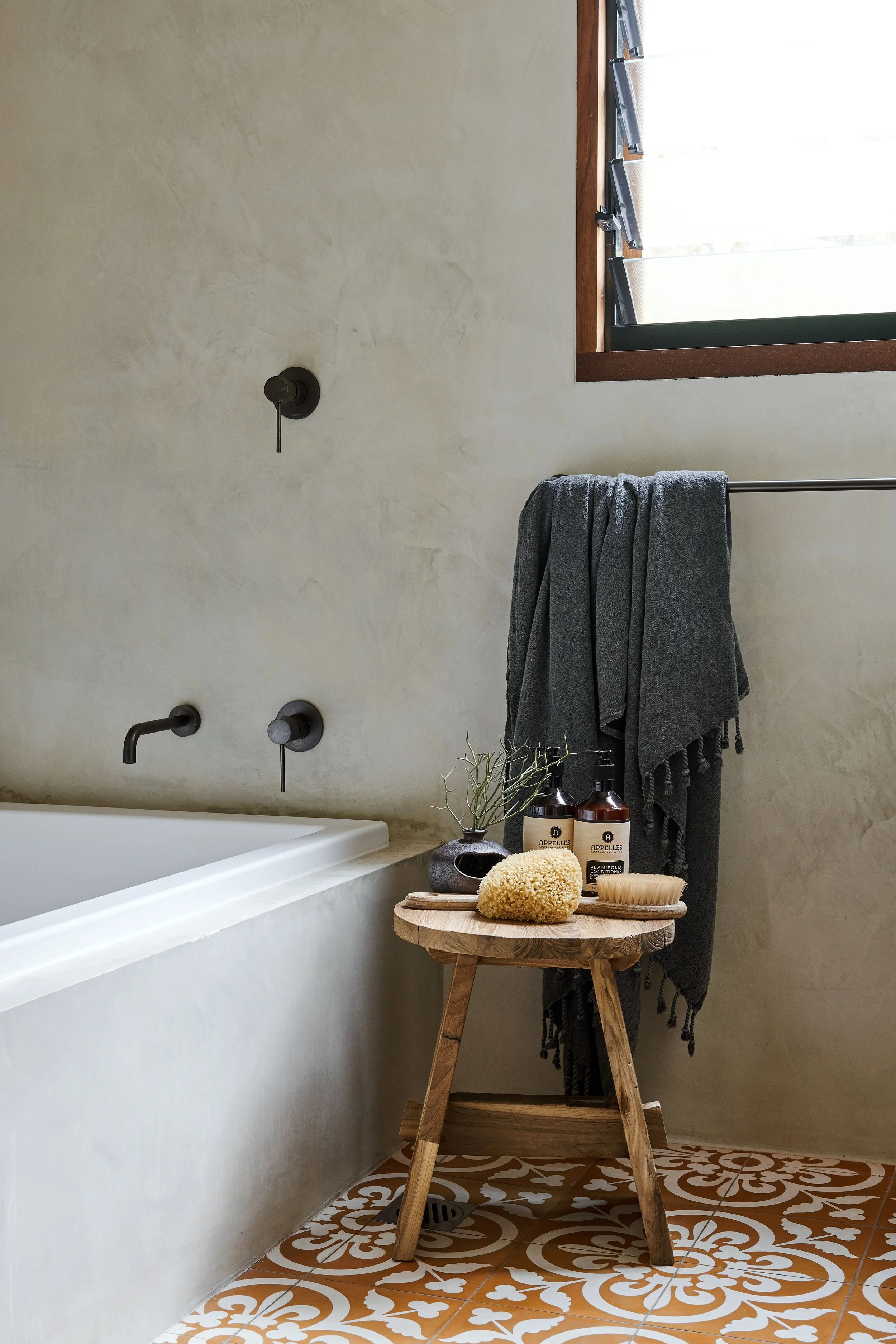 Bathroom with a bathtub, a small wooden stool with bath products, a gray towel, a potted plant, and decorative patterned floor tiles.