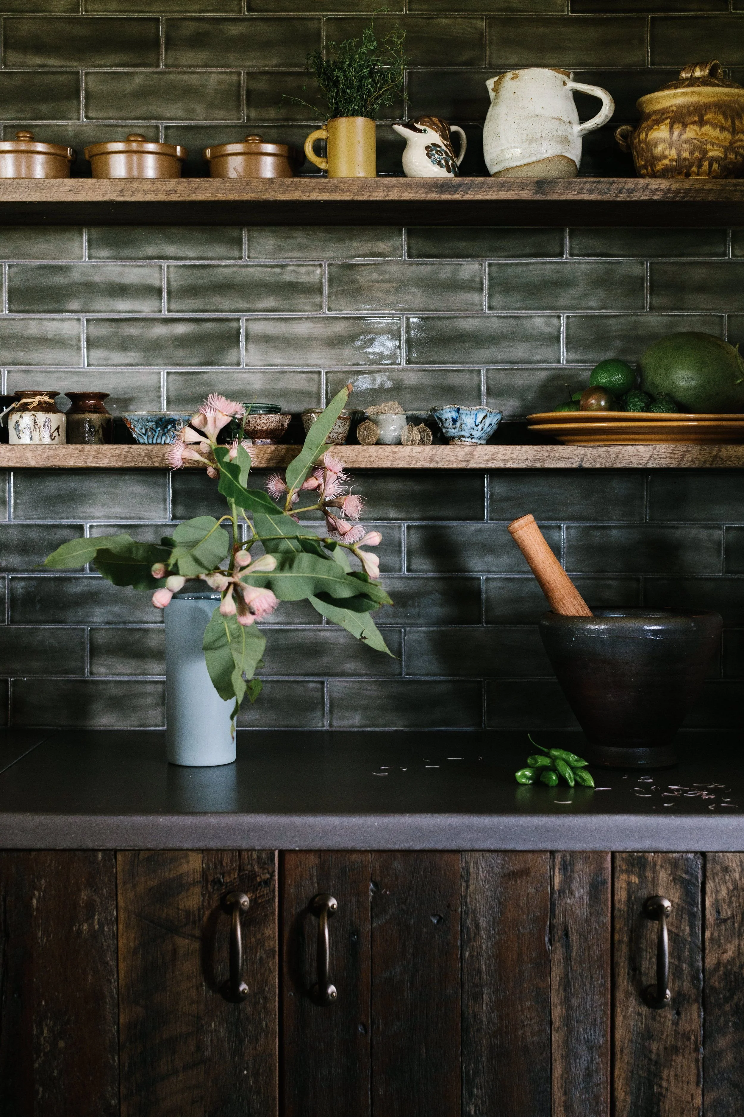 A kitchen counter with a vase holding pink flowers and green leaves, a mortar and pestle, and small scattered pink petals.