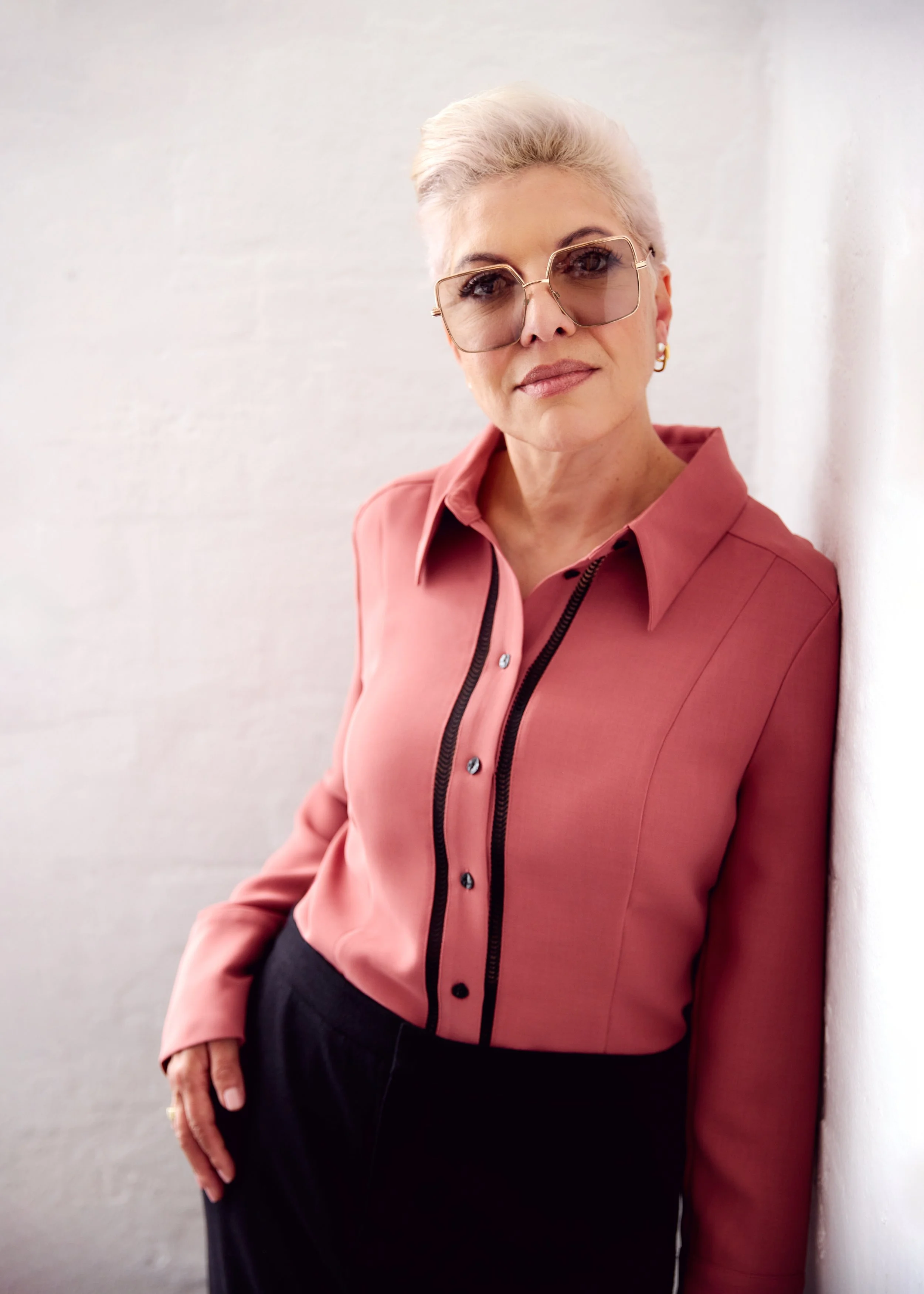 A mature woman with short, platinum blonde hair wearing sunglasses, a pink button-up shirt, and black pants, standing against a white wall.