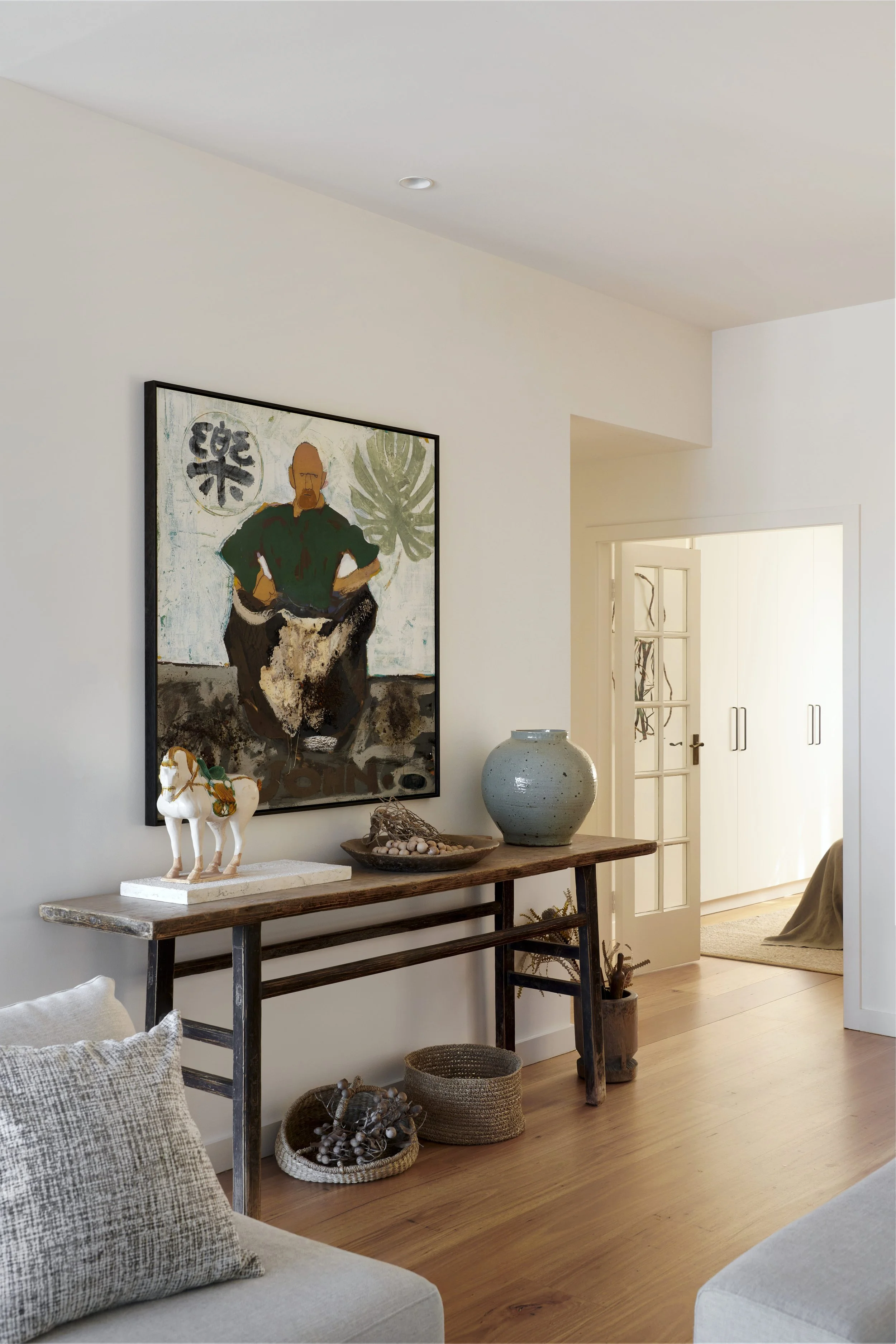 Interior of a living room with a console table holding decorative items, hanging painting, and light-colored walls and flooring.
