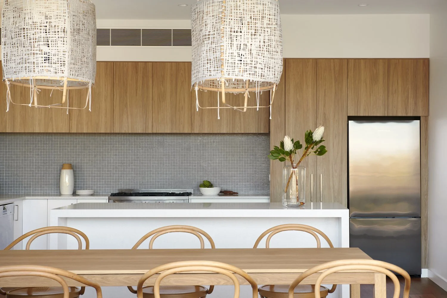 Modern kitchen with wooden cabinets, gray tile backsplash, white countertops, stainless steel refrigerator, and a wooden dining table with matching chairs. Hanging woven pendant lights and a vase with white flowers on the counter.