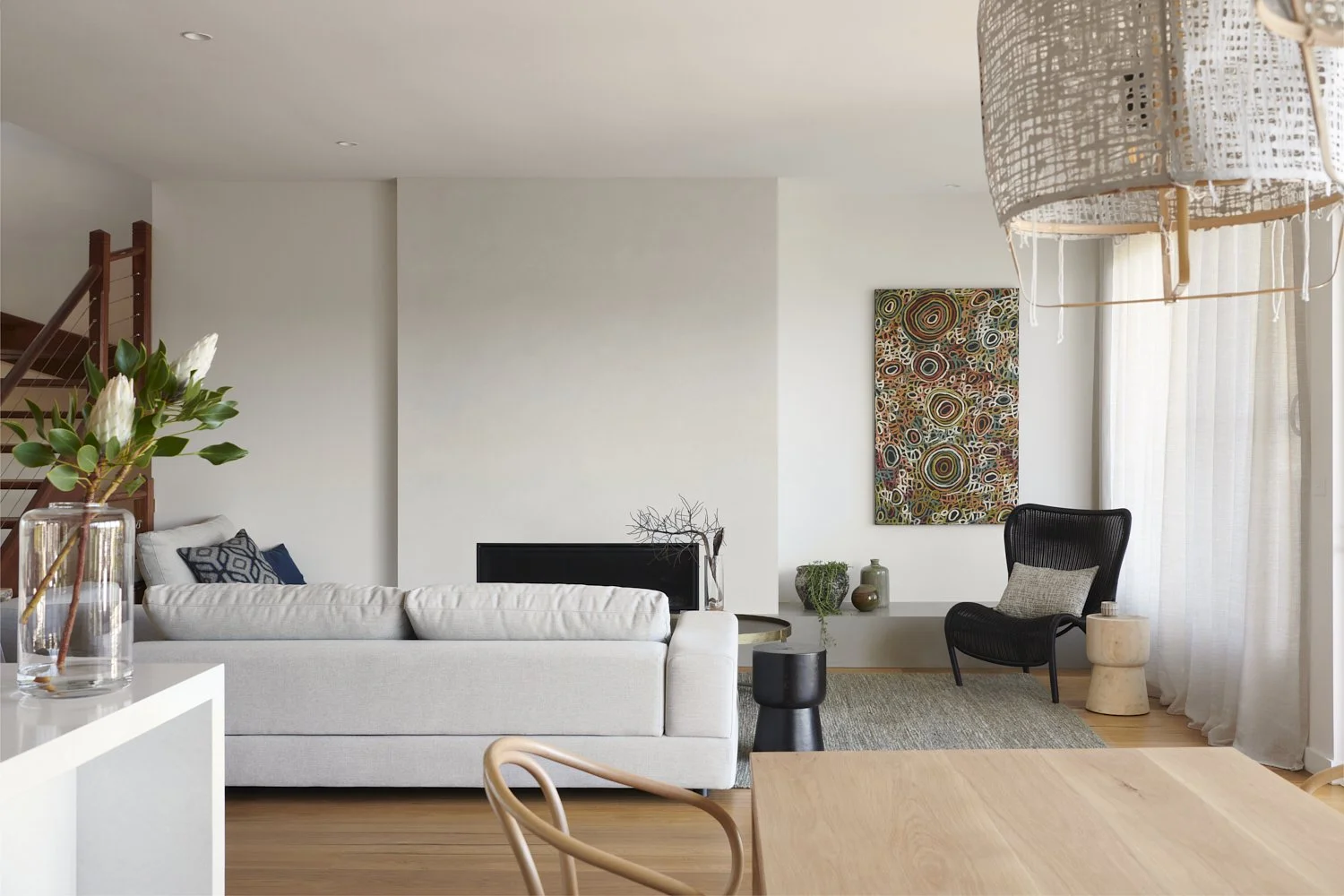 Modern living room with white sofa, black armchair, colorful abstract art on wall, floor-to-ceiling sheer curtains, wooden furniture, decorative vases, and greenery.