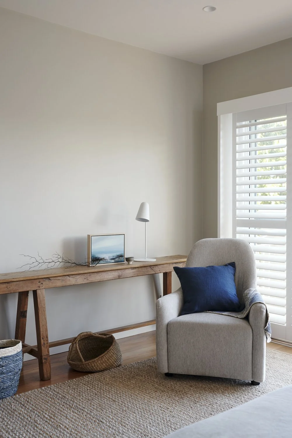 A cozy corner of a living room with a beige armchair, a dark blue pillow, a wooden console table with a small lamp, a seashell picture, and woven baskets, next to a window with white shutters.