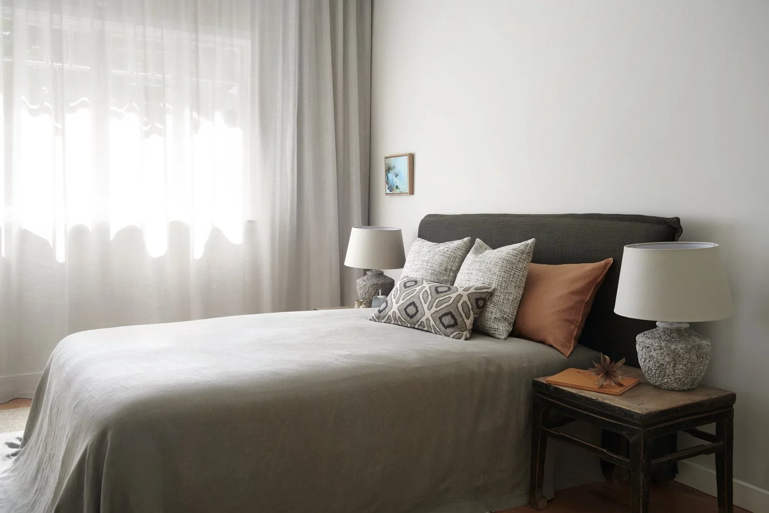 A bedroom with a bed, lamps, pillows, a window with curtains, a small painting, and a wooden side table with decor.