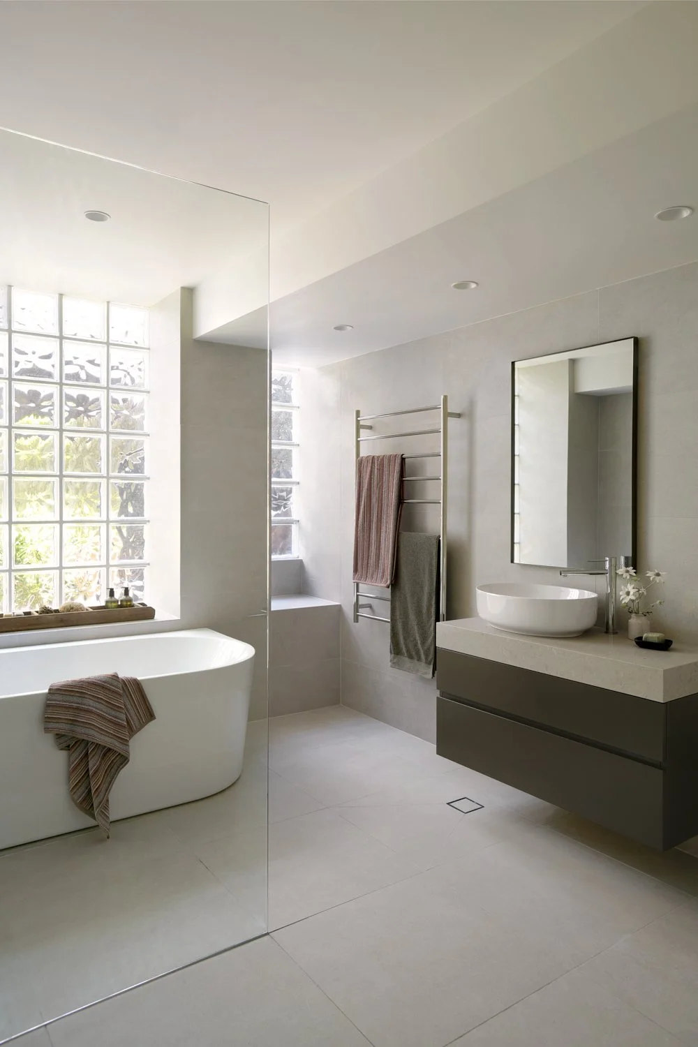 Modern bathroom with a white bathtub, a mirror, a vessel sink, a towel warmer with towels, and a glass block window allowing natural light.