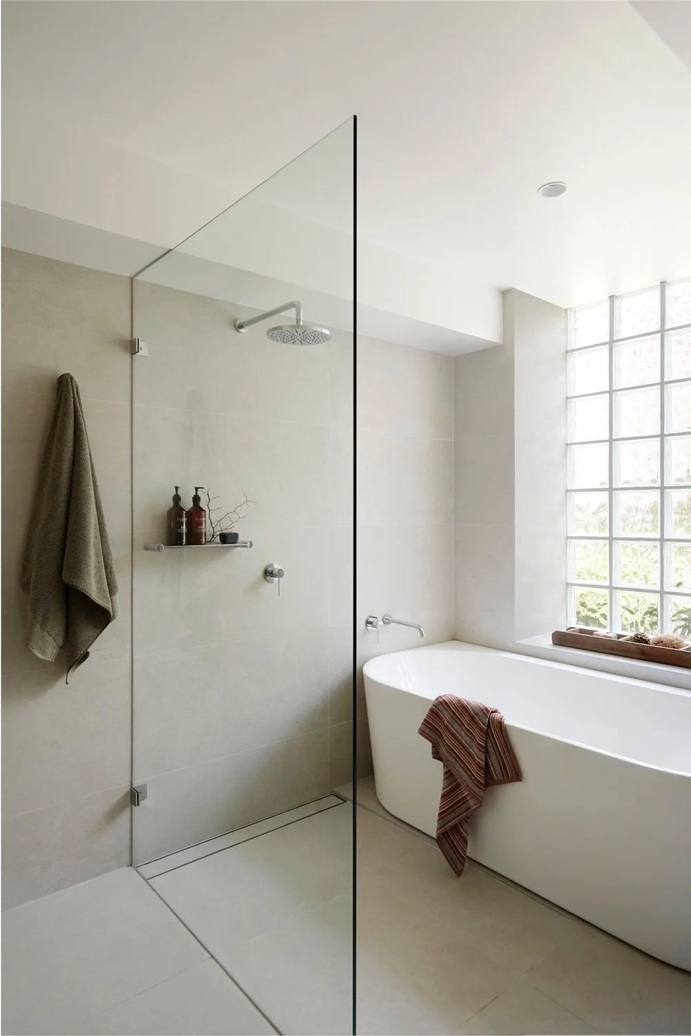 Minimalist bathroom with a glass partition, walk-in shower, beige tiles, modern bathtub, large window with grid pattern, bathroom towels, and toiletries on a shelf