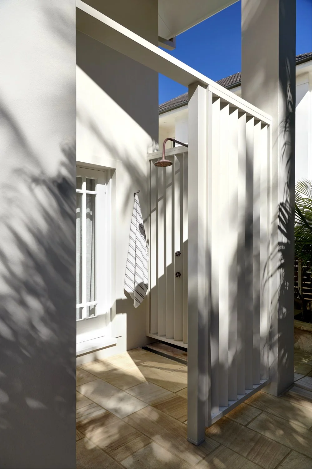 Shower area outdoors with white walls and a white door with bars, a striped towel hanging, a wall-mounted showerhead, and shadows cast by nearby plants.