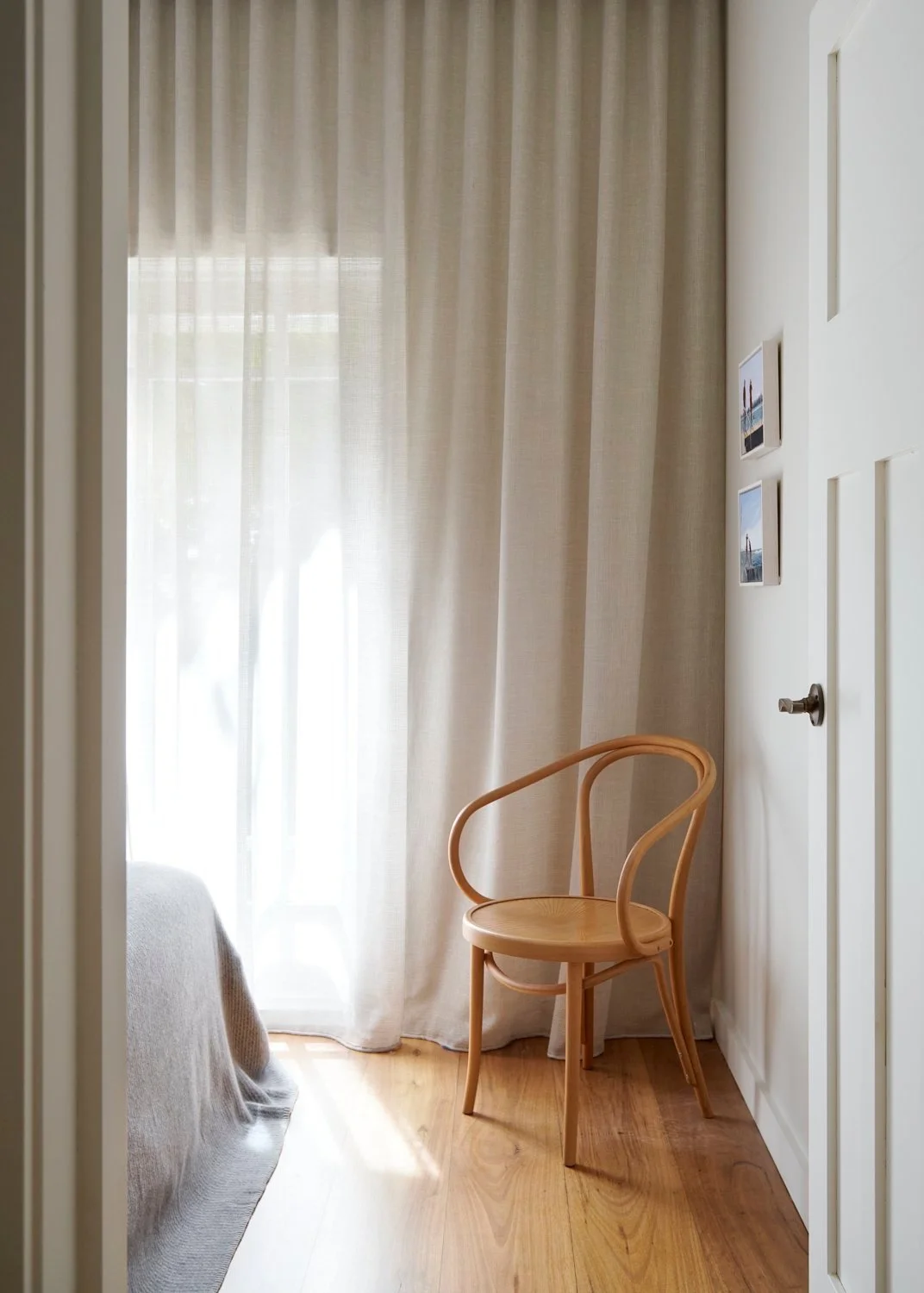 A cozy corner with a wooden chair next to a window with sheer curtains, sunlight streaming through, and framed photographs on the wall.