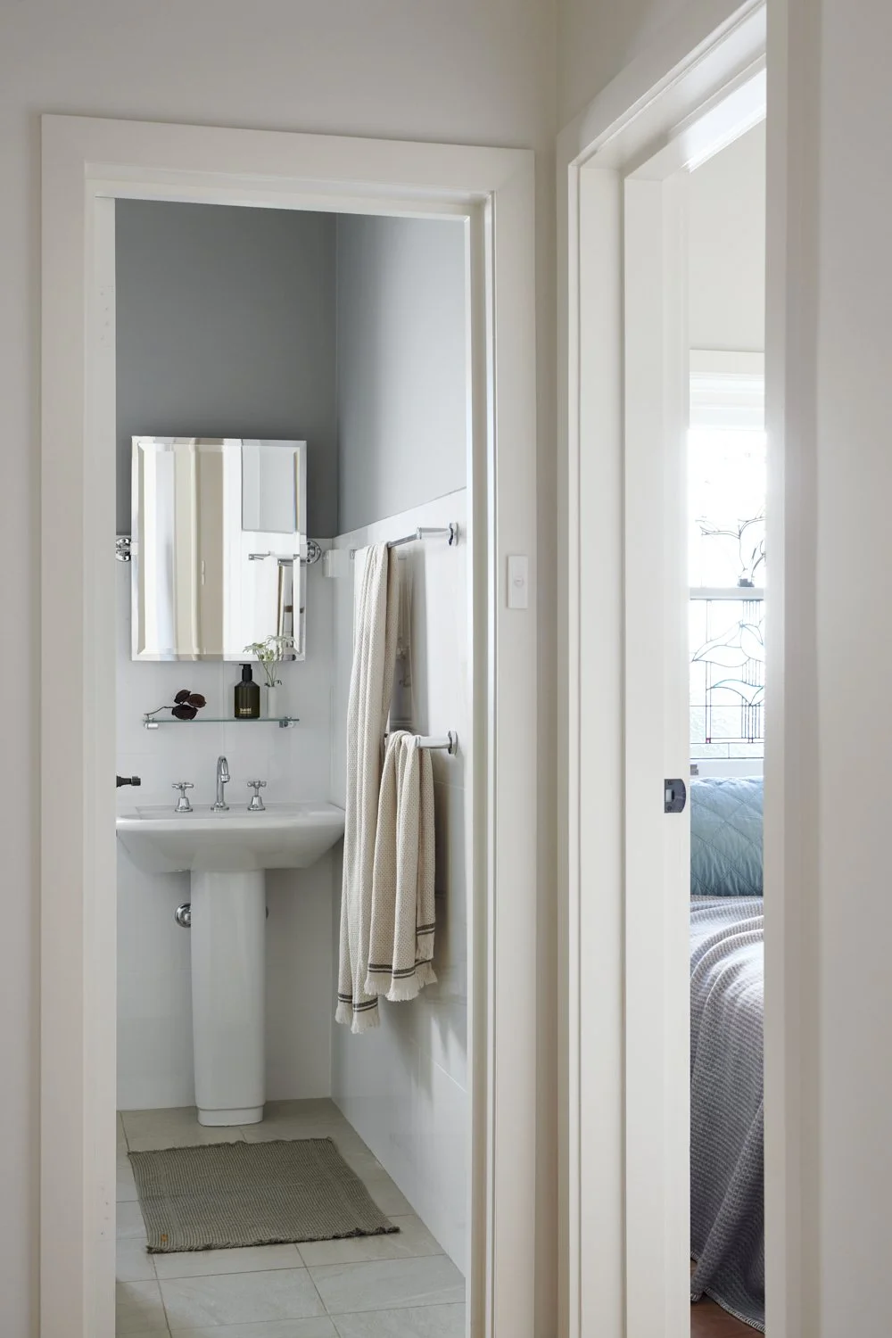 A small bathroom with a pedestal sink, a small mirror, and a glass shelf with decorative items. The bathroom has gray and white walls, with a towel rail holding two beige towels. There is a beige rug on the floor. To the right, there is an open door leading to a bedroom with a bed and a large window with decorative wrought ironwork.