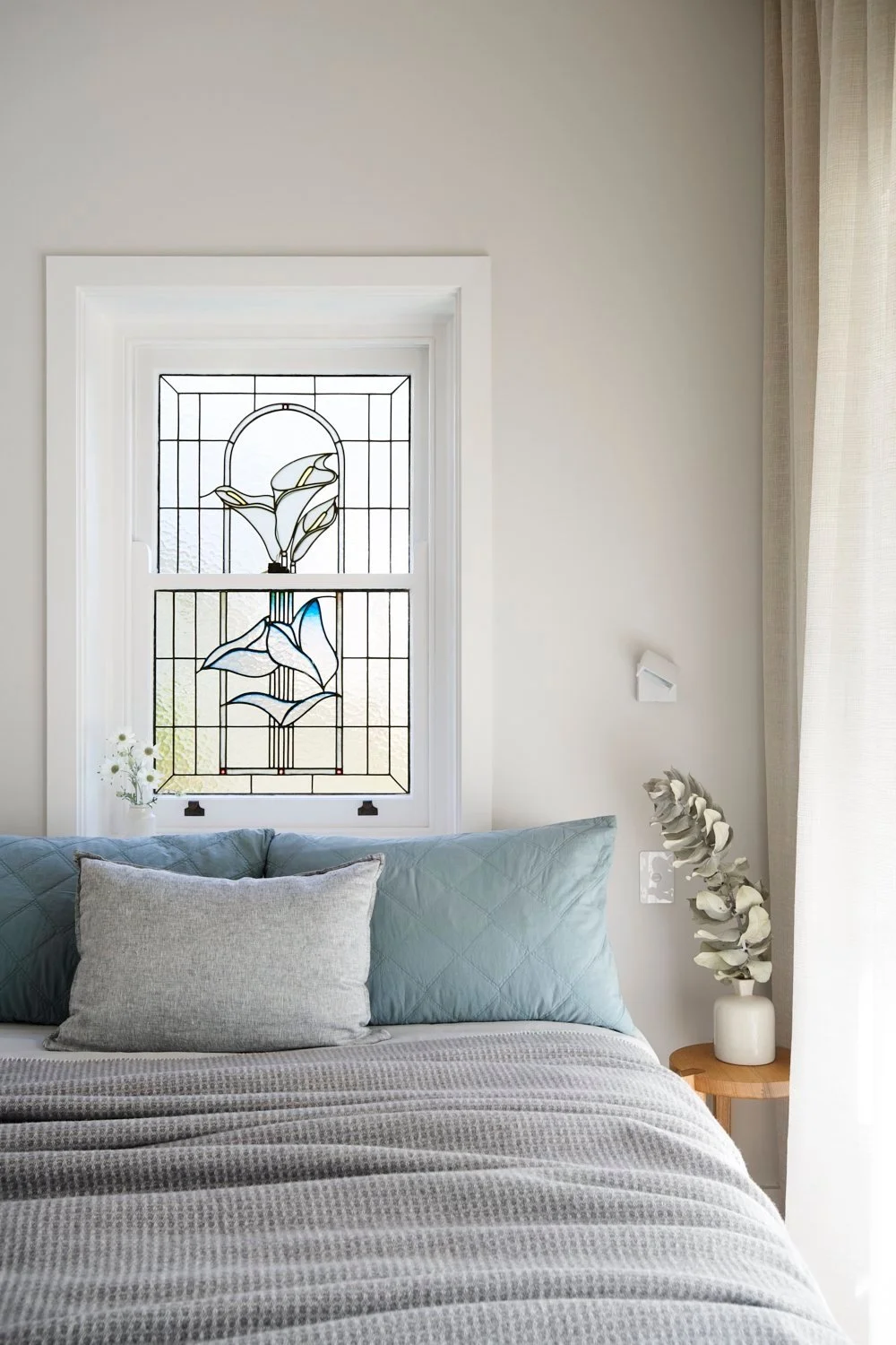A bedroom with a stained glass window above the bed, white walls, a gray bedspread, blue pillows, a small wooden side table with white flowers, and light-colored curtains on the right.