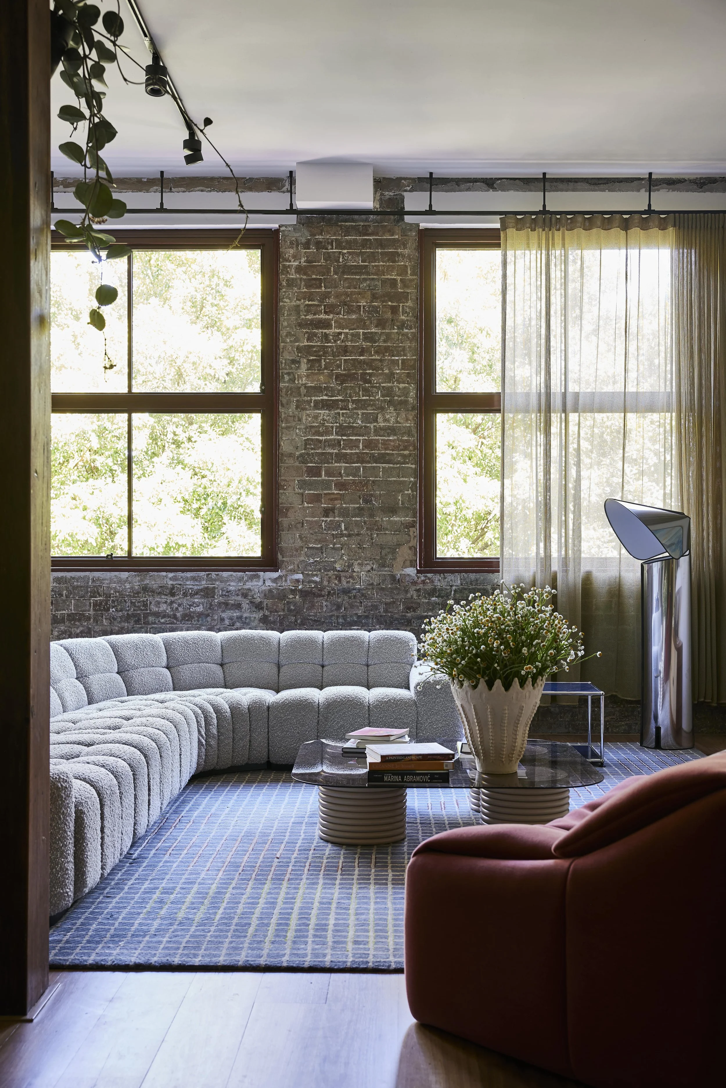 Living room with exposed brick wall, large windows, gray curved sofa, purple armchair, glass coffee table with books and a large vase of white flowers, and sheer curtains.