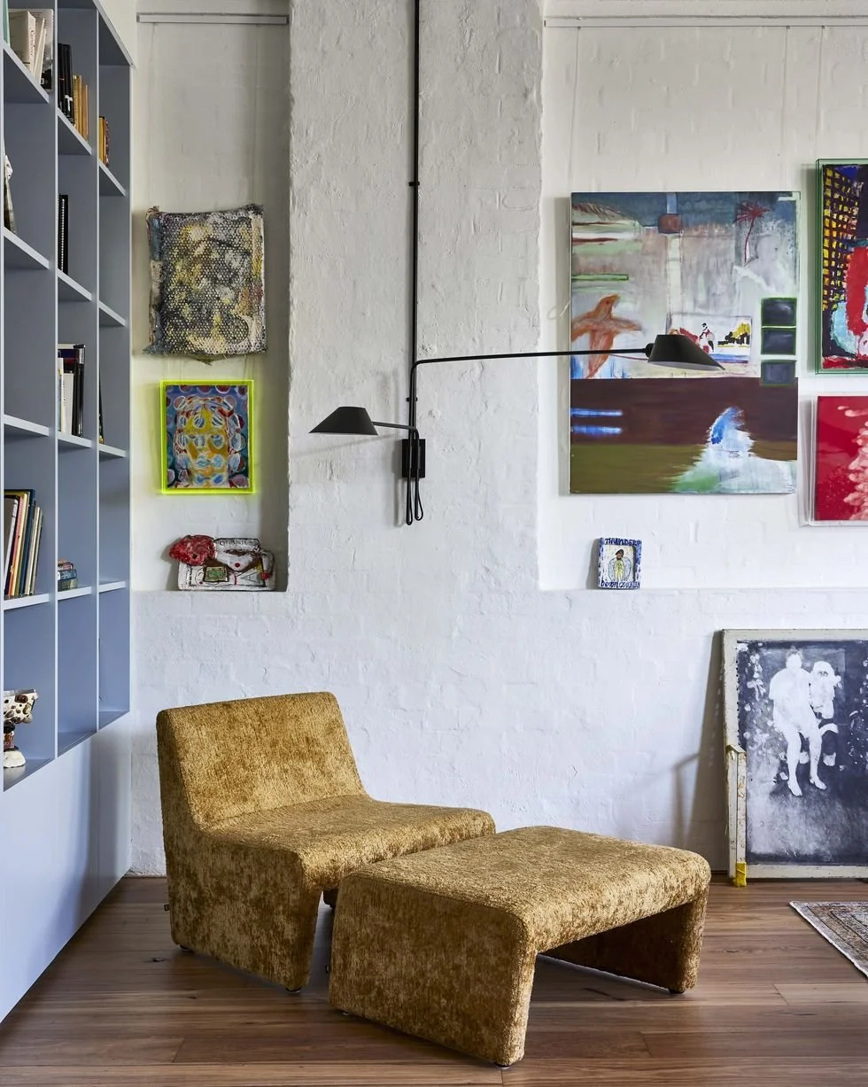 Interior of an art gallery with a mustard-colored lounge chair, white brick wall decorated with abstract paintings, and a gray bookshelf filled with books.