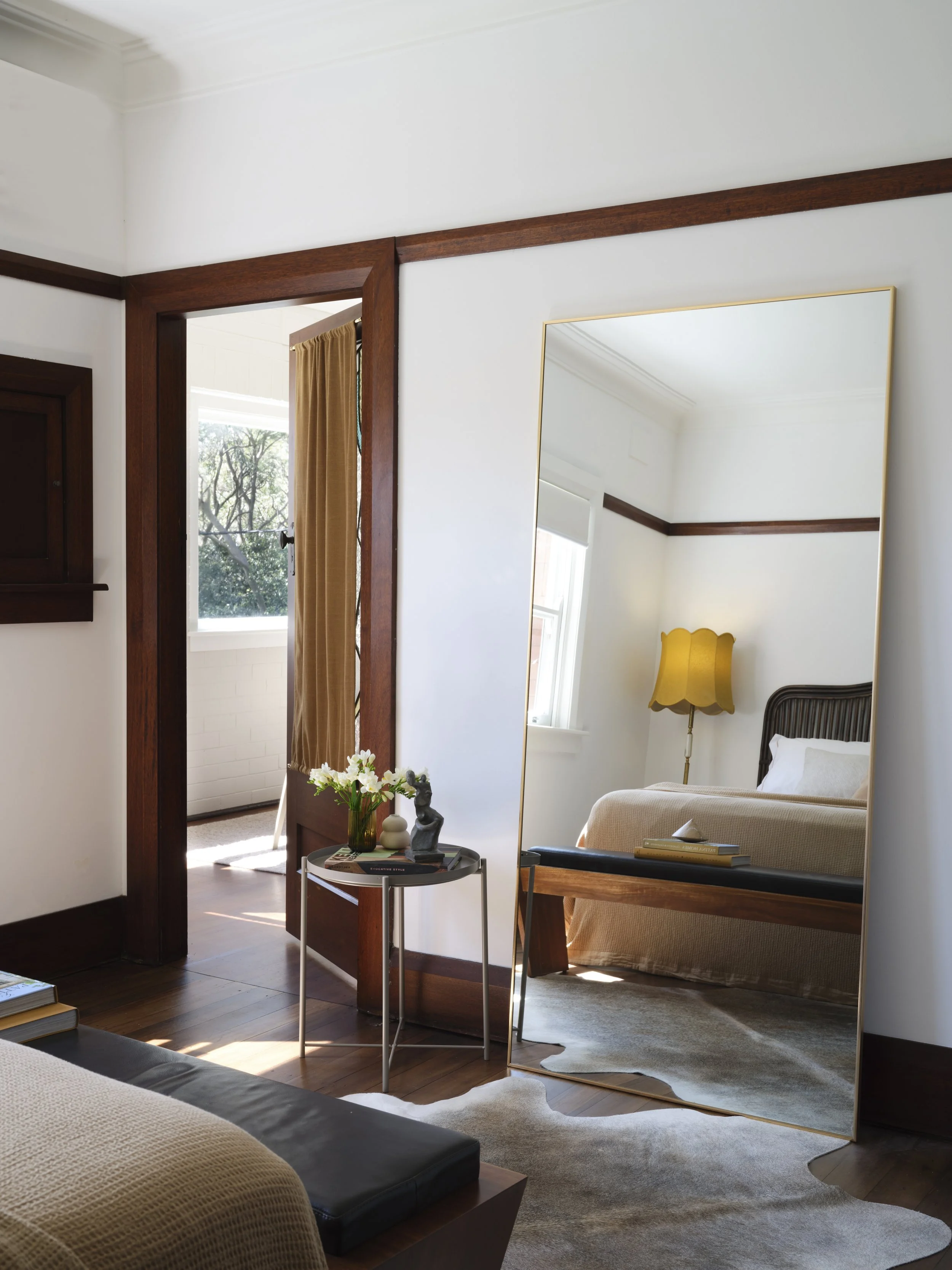 Interior of a bedroom with a large mirror reflecting the bed, a yellow lampshade, and a window with sunlight. A small table with decorative items and flowers is beside the bed. The room has wooden trim and a light-colored rug.