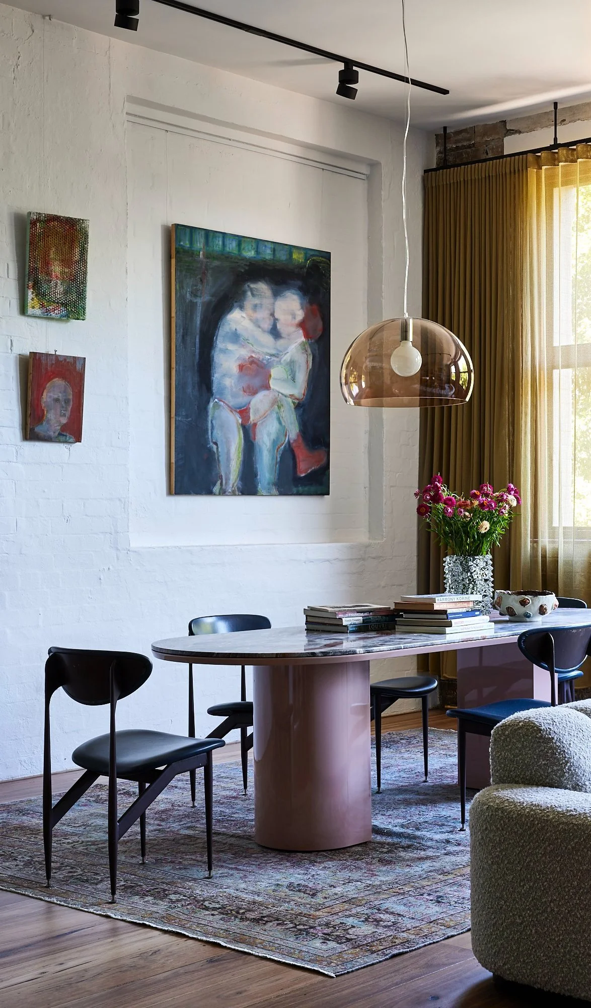 Living room with white brick wall, colorful artwork, a dining table with books and a vase of flowers, black chairs, a patterned rug, and a window with yellow curtains.