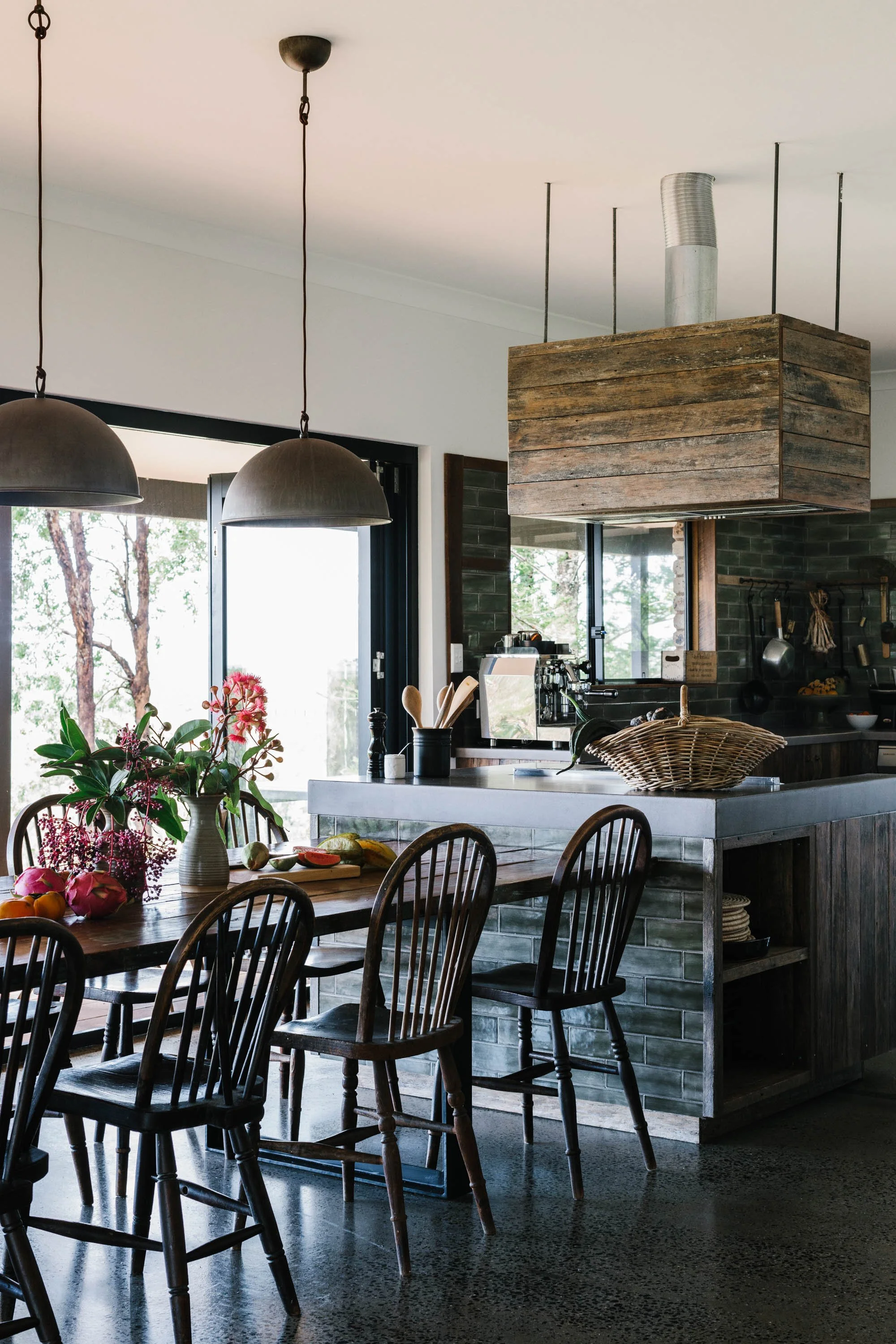 A cozy kitchen and dining area with wooden chairs and a table decorated with flowers and fruits. Modern pendant lights hang from the ceiling. The kitchen features a gray countertop, a basket, and various cooking utensils. Large windows bring in natural light and view of trees outside.