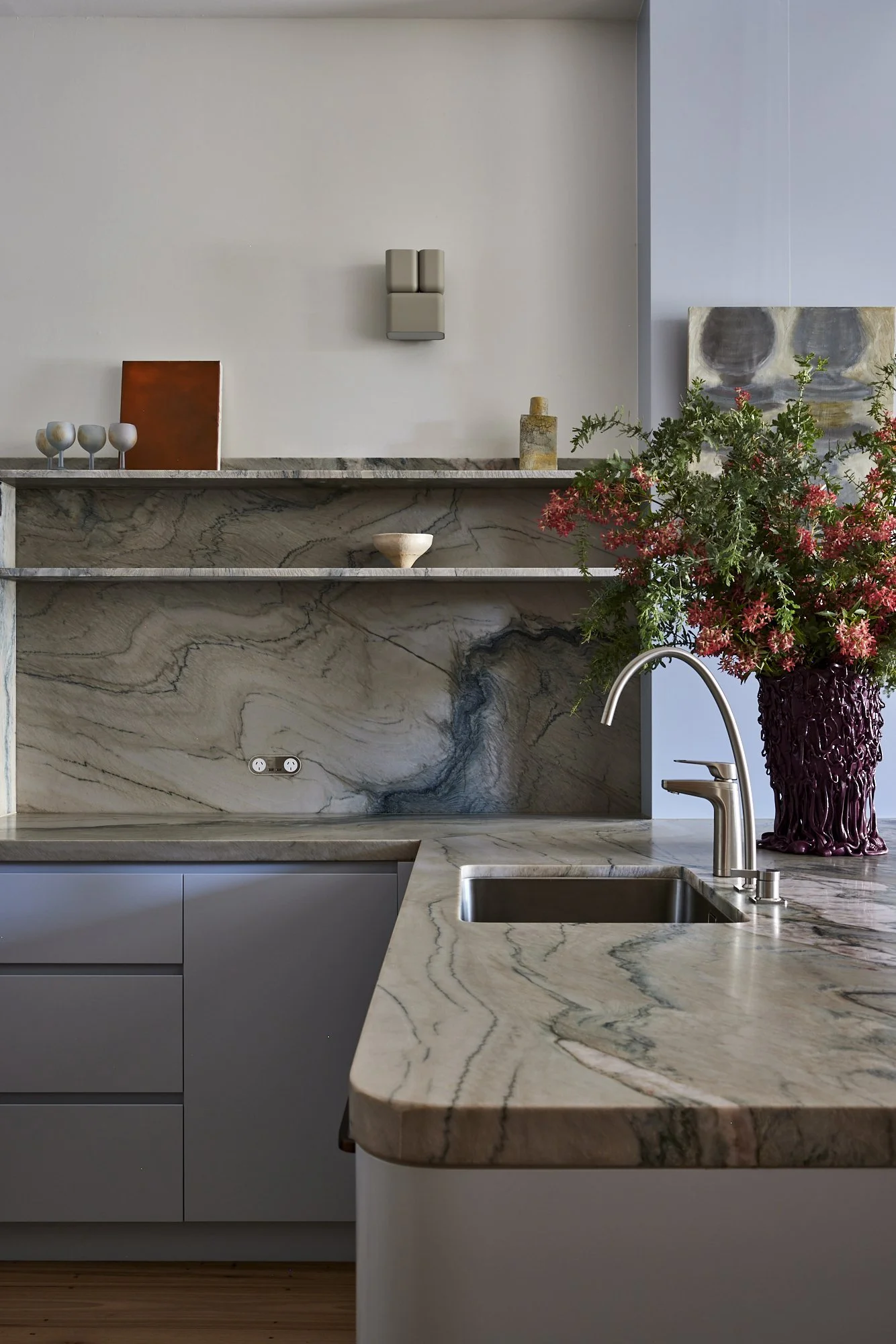 Modern kitchen with a marble countertop, a stainless steel sink with a curved faucet, and a bouquet of pink flowers in a dark purple vase.