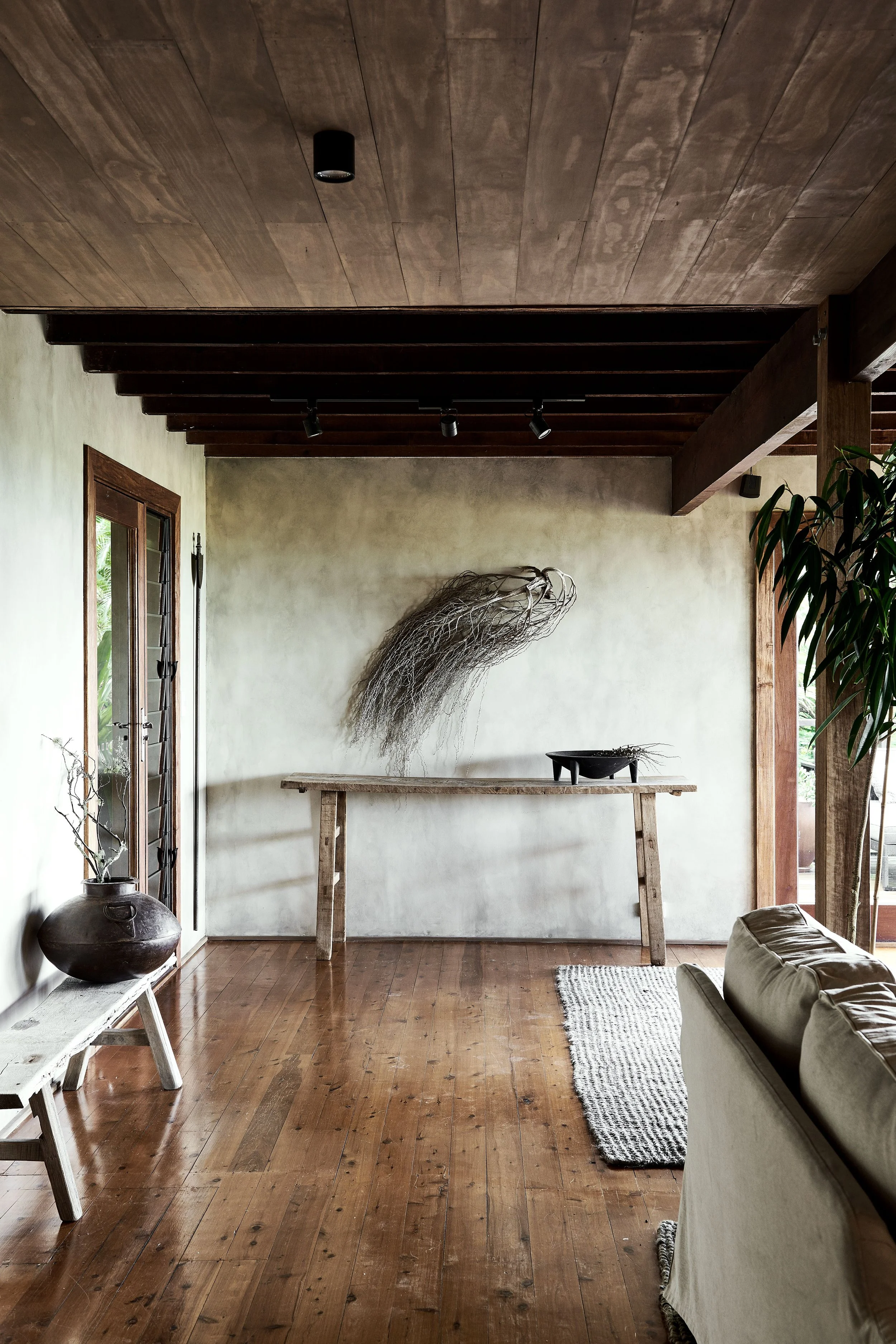 Interior of a room with wooden floors and ceiling, a sofa, a table against the wall decorated with dried plants or coral, and a large potted plant near the window.